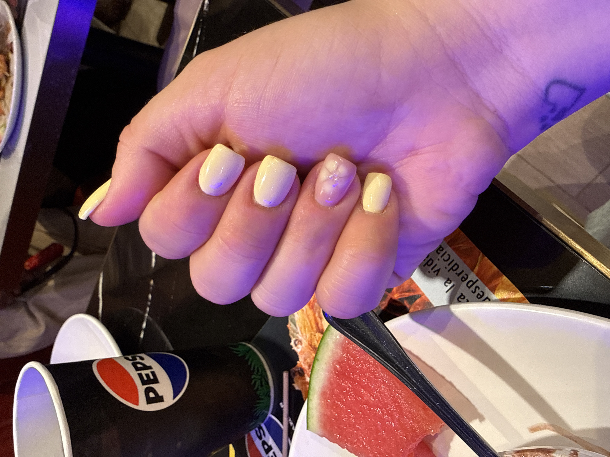 A person's hand, adorned with pastel yellow and light purple nails, is the central subject of this media file. One nail, on the ring finger, has a subtle floral design in white and pink. The hand is positioned over a collection of objects and a partially visible scene.
In the foreground, a black disposable cup with the red, white, and blue Pepsi logo is visible. Next to it, a slice of watermelon is partially submerged in a white bowl, with a black utensil resting on the rim of the bowl and partially covering the watermelon. In the background, other objects are out of focus, including what appears to be a white plate on the left and dark surfaces.
The lighting suggests an indoor setting, possibly a restaurant or cafe, with a purple hue cast over the hand, potentially from ambient lighting or a nearby light source. The overall mood conveyed by the image is casual and relaxed. The visible text is limited to the Pepsi logo on the cup. The provided location context is Carretera De San Martín, Spain, which suggests this scene might be taking place in that area. The time of day and weather are not discernible from the image.