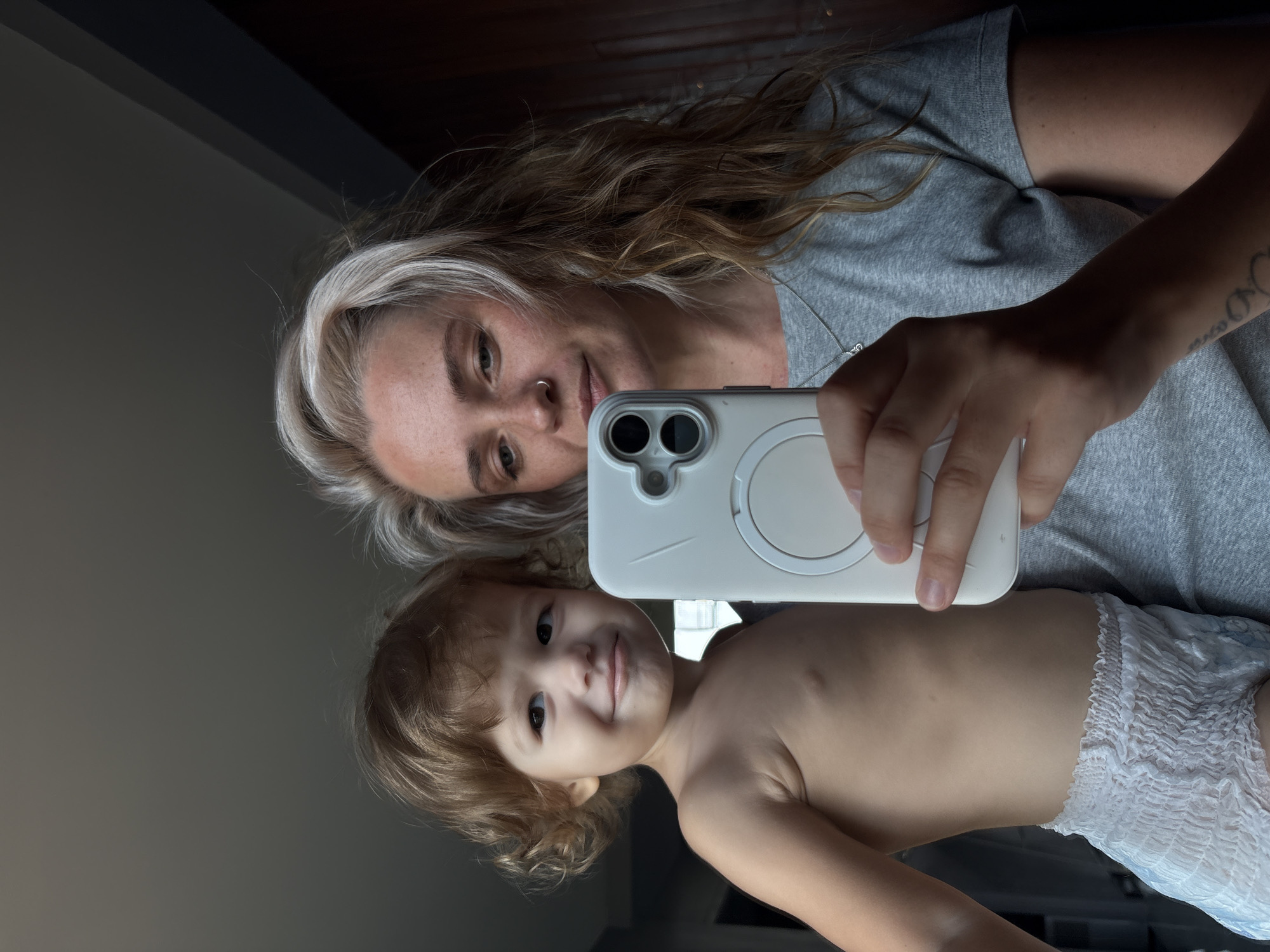 This image is a mirror selfie featuring an adult woman and a young child, likely a mother and child. The image is correctly oriented horizontally for viewing.
The woman is positioned towards the upper left of the frame. She has long, wavy brown hair with prominent blonde highlights, a nose piercing, and a fair complexion. She is wearing a simple grey short-sleeved t-shirt and a delicate silver necklace with a circular charm. A tattoo is visible on her left forearm. She is holding a white smartphone with a matching white case, which features a distinctive circular magnetic attachment (likely MagSafe compatible) on its back. Her gaze is directed towards the camera/mirror, and she has a gentle smile.
Below and slightly to the right of the woman is the young child, who appears to be a toddler. The child has light brown, curly hair and dark eyes, looking directly at the camera/mirror with a slight, innocent smile. They are bare-chested and wearing a white, disposable pull-up diaper.
The background reflected in the mirror is dark and indistinct, suggesting an indoor setting. Both individuals appear calm and content. No visible text is present in the image.