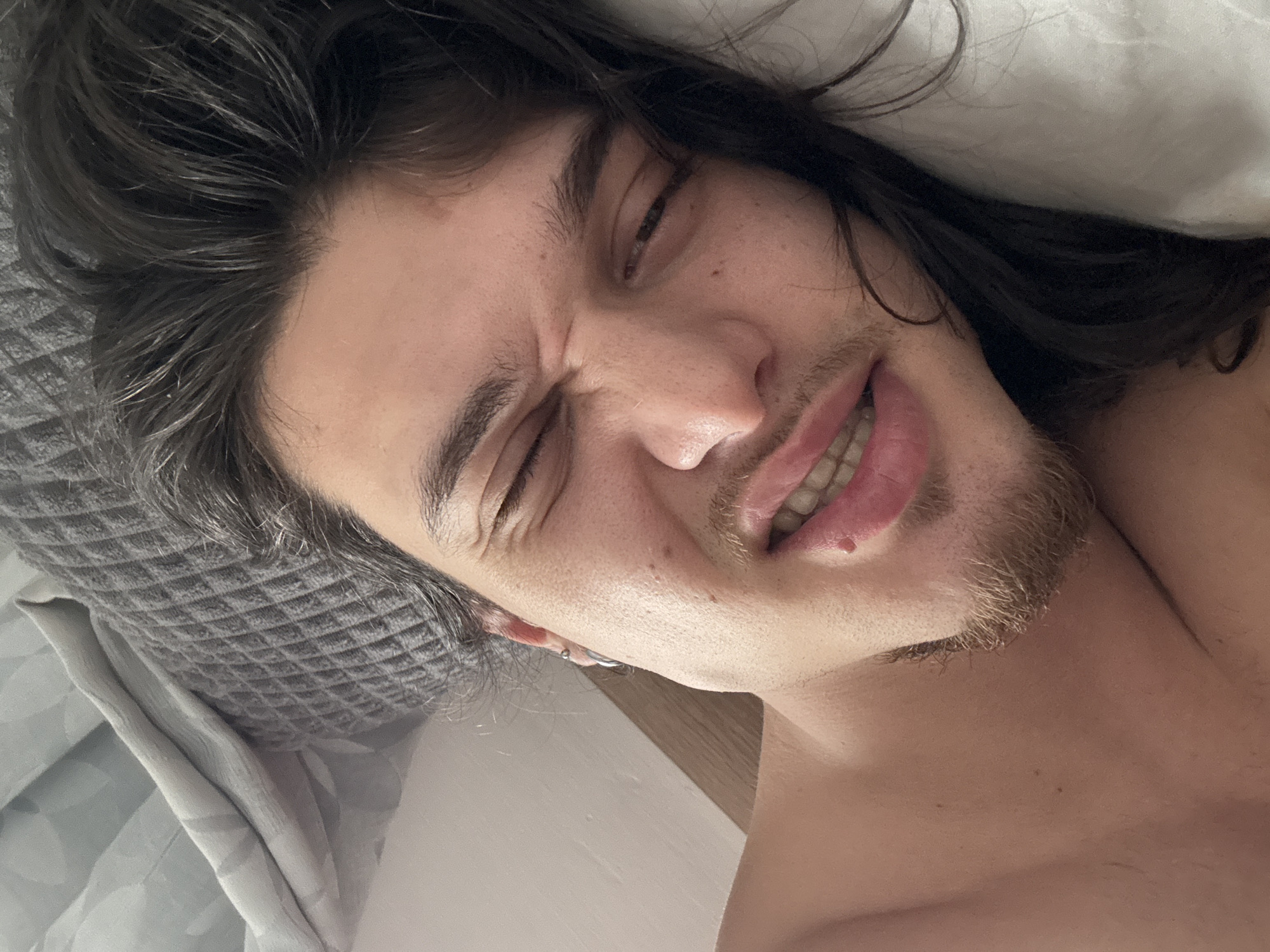 A close-up, angled, personal photograph depicts a light-skinned male in apparent discomfort. He lies on a gray pillow and white bedding. His eyes are closed, and his face is contorted in a grimace, revealing his teeth. He has dark, shoulder-length hair and a light beard. No specific information ties the image to Sofia, Bulgaria. No specific location or activity can be identified.