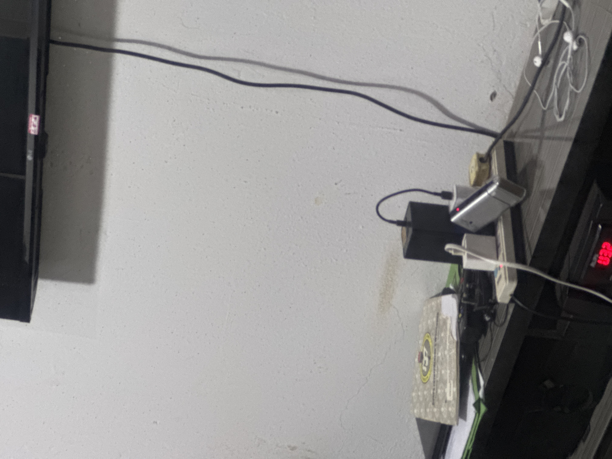 This media file shows a close-up of electrical cords and charging devices plugged into a wall outlet. The scene is likely indoors, given the presence of electronics and what appears to be a textured wall. Two cables, one black and one gray, run horizontally across the frame, casting shadows on the wall. In the middle of the image, a power strip is visible, with several items plugged into it. These include a black power adapter, a silver device that appears to be a portable charger or battery pack with a red indicator light, and a white adapter. A white cable with an earbud-like attachment is also present, suggesting the use of headphones. To the right, a black shelf or desk is visible, holding more electronic equipment, including a device with a red digital display that shows the text "32". A portion of a box with a yellow and black circular logo is also visible. The lighting suggests it could be daytime, but the overall impression is one of a cluttered workspace or entertainment area. No people are visible. The focus is on the arrangement of wires and devices, indicating a potential need to manage or organize these electronics.