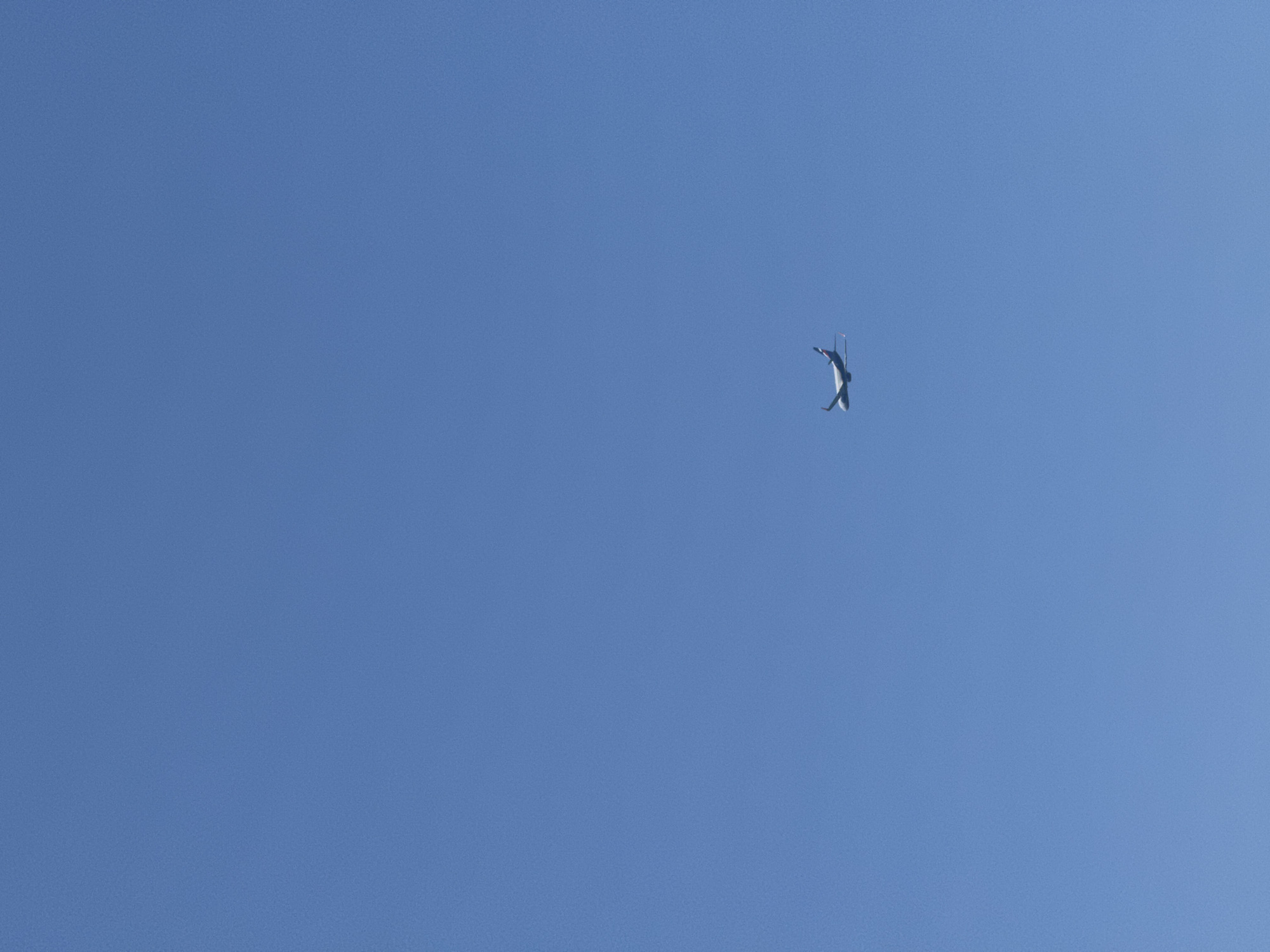 A single airplane is visible in a clear blue sky. The airplane is white with blue and red accents on its tail. It appears to be flying at a high altitude. The image is taken from a ground-level perspective, looking up at the sky. The location is stated to be Sochi, Russia.