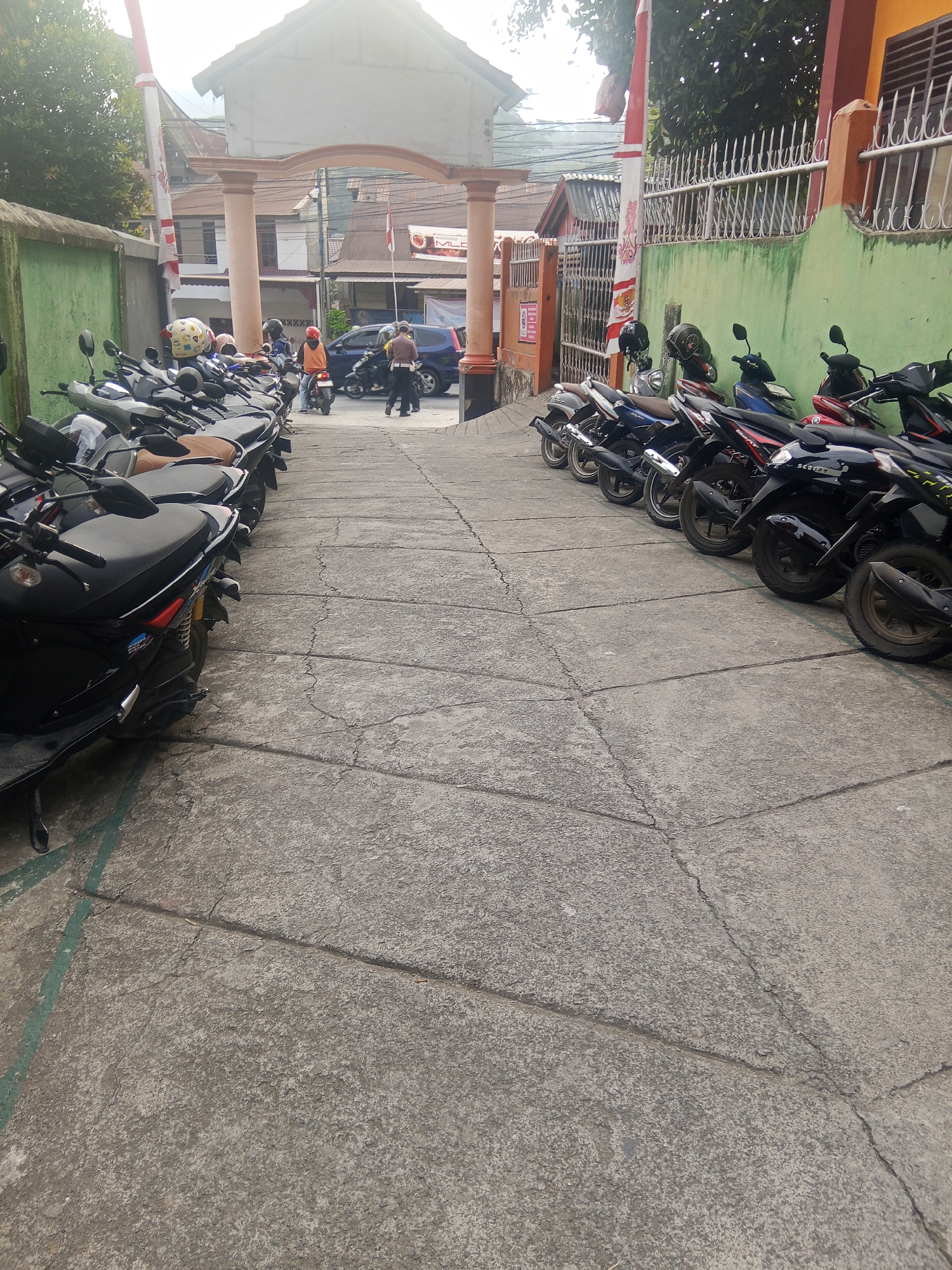 The image depicts a parking area for motorcycles in Makale, Indonesia, likely near a public building or facility. In the foreground, a row of parked scooters is visible on a concrete surface that is cracked and weathered. The scooters are predominantly dark-colored, with some featuring brown seats. A green line is painted on the concrete to the left, indicating a parking boundary.
In the background, an arched gateway with pillars frames the scene. Beyond the gateway, several people are present. One individual, wearing an orange jacket and a helmet, is seated on a scooter. Another person in a brown uniform appears to be directing traffic or observing the area. A blue SUV is also visible behind the scooters.
The environment suggests a sunny day with some overcast conditions, as indicated by the lighting. Trees are visible to the left and right, and buildings with tiled roofs are present in the distance. The overall impression is of a functional, everyday scene in an Indonesian urban setting.
