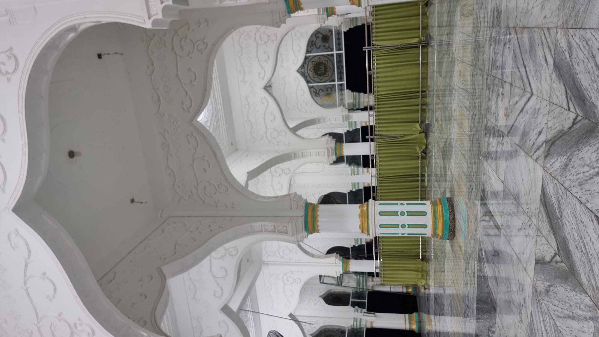 An interior shot shows a mosque with white arched ceilings and columns, featuring intricate white decorative carvings. The floor is made of polished grey and white marble tiles, reflecting the overhead lights. A section of the mosque interior is separated by a line of green curtains draped from silver poles. A prominent white and gold column with green accents stands in the foreground, positioned centrally. In the background, through the arches, other columns and green curtains are visible, along with a decorative arched window featuring a circular emblem. The scene appears to be located in Lapang Timu, Indonesia, and is devoid of people.