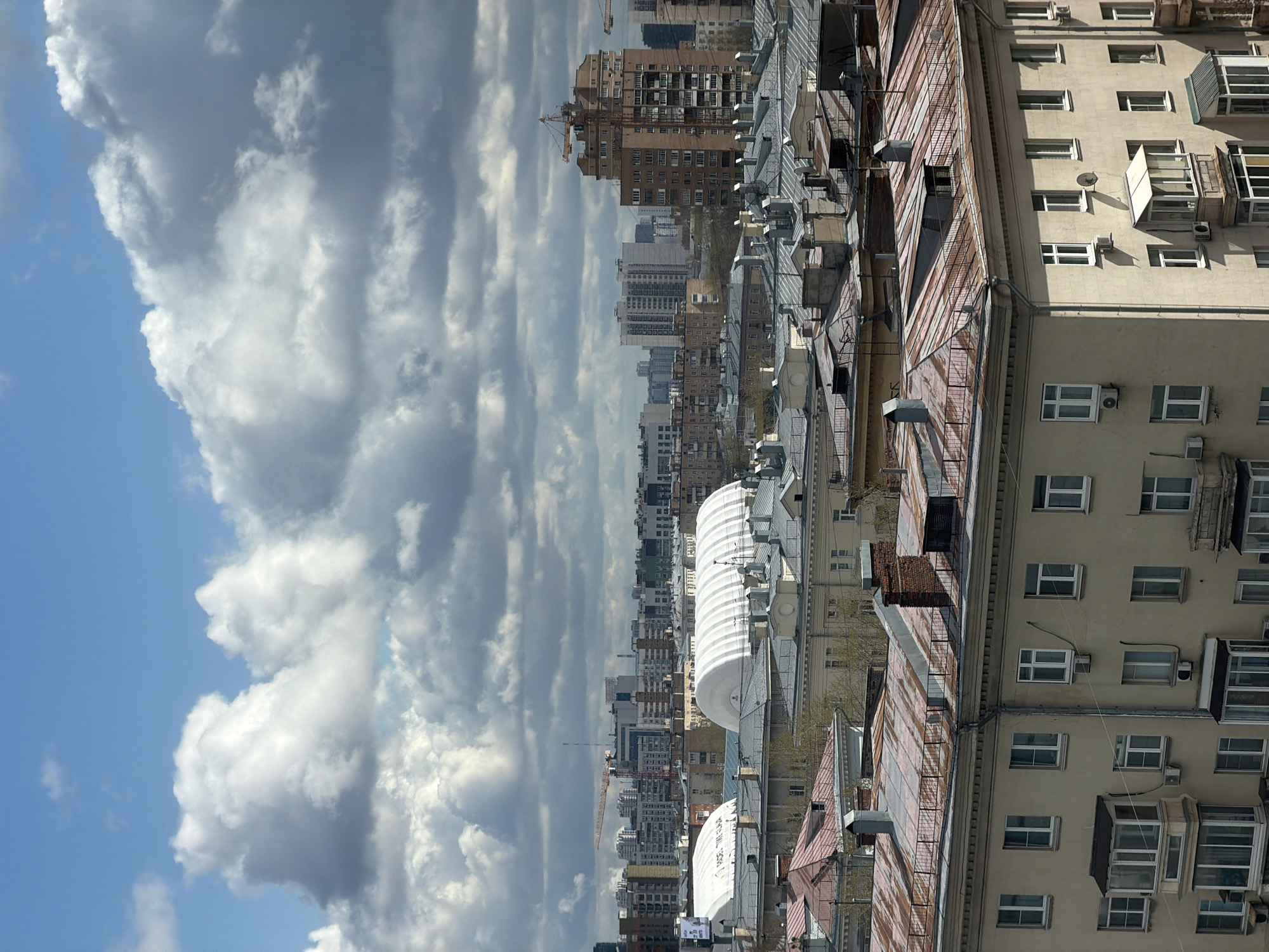 The image shows a high-angle view of Moscow, Russia. The sky on the left is partly cloudy with white and grey clouds against a blue sky. The cityscape includes various buildings, ranging from apartment buildings to taller office blocks. A construction crane is visible amongst the buildings. In the foreground are rooftops of buildings with air conditioning units and windows. Some buildings have a light beige exterior.