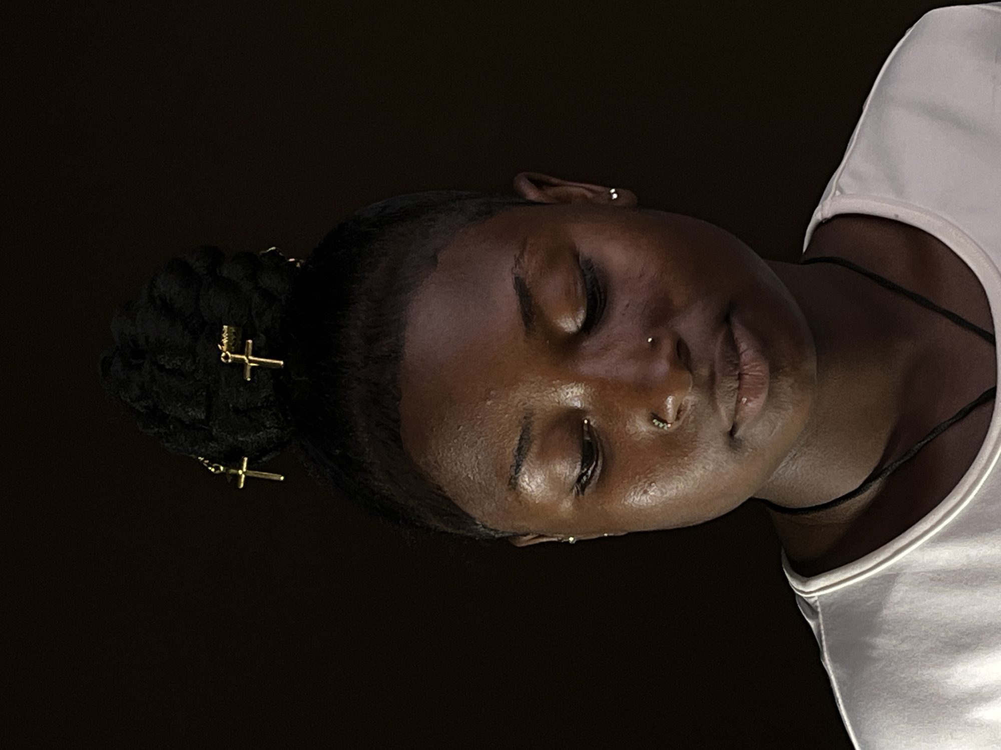 The image features a person with dark skin, positioned with their head tilted to the right and eyes closed, suggesting a state of rest or calm. Their dark hair is styled into a braided bun, embellished with multiple gold-colored, cross-shaped hair accessories. The person has a small stud piercing on their left nostril and a septum ring. They are wearing a white t-shirt or tank top, and a thin black cord is visible around their neck. The background is a solid, dark tone, indicating low lighting, possibly nighttime or an indoor setting. The scene evokes a sense of peace and tranquility. This image was captured in Barakin Akawo, Nigeria. There is no visible text.