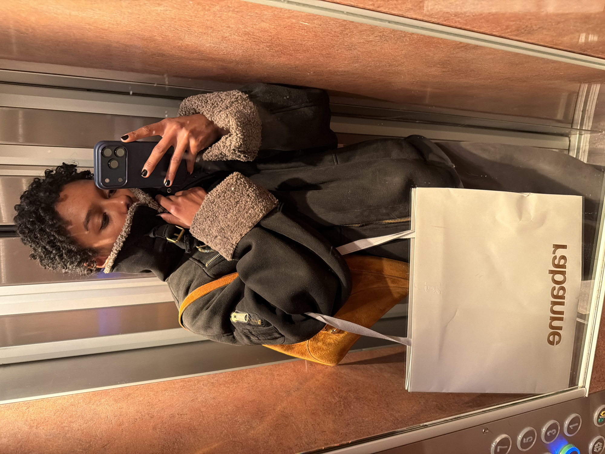 A person takes a selfie in an elevator. The person has black, curly hair and is wearing a black jacket with shearling trim. They are holding a smartphone with a blue case. There is a white shopping bag with the word "Sapsue" printed on it in reverse in the elevator. The elevator has a mirrored wall and orange walls and a stainless steel control panel. It is highly probable that the shopping bag indicates that the location is Paris, France as "Sapsue" is "Capsule" reversed and Capsule is a clothing store located in Paris.