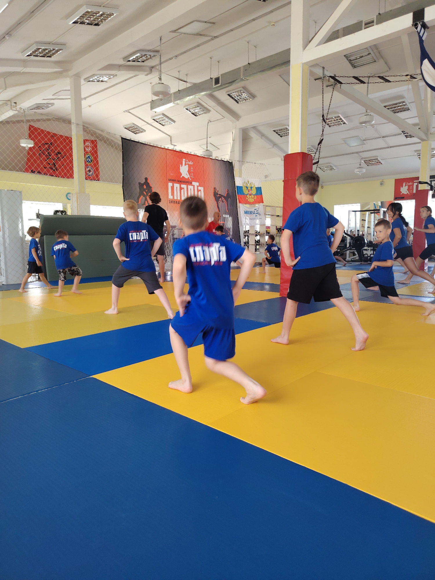 In a gymnasium with a blue and yellow mat floor, a group of children in blue shirts and black shorts are performing exercises. The gym has a high ceiling with fluorescent lighting and exposed beams. Red banners with black graphics are hung on the walls, one featuring silhouettes of people engaged in martial arts and another displaying the Russian flag. Several children are in motion, with one boy in the foreground extending his left leg back in a lunge. Other children are visible in the background, also engaged in physical activity. The scene appears to be a martial arts training session for young athletes, possibly at a sports school or club in Chaikovsky, Russia, indicated by text on the banners that includes "СПАРТА" and "ЗАПАД".