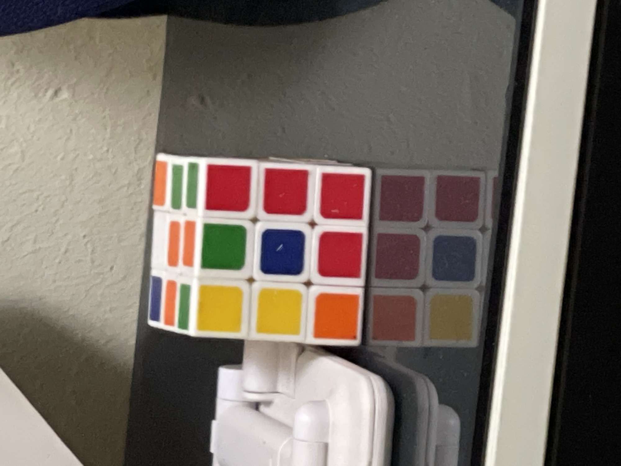 A Rubik's cube is mounted to a reflective surface, likely a glass panel or door, using a white plastic mount. The cube's visible faces display red, white, orange, yellow, green, and blue squares. The reflective surface shows a blurred reflection of the cube. The background includes a textured wall and a dark-colored surface above the cube. The scene is indoors. No information can be derived from the image that identifies the location as Abuja, Nigeria.