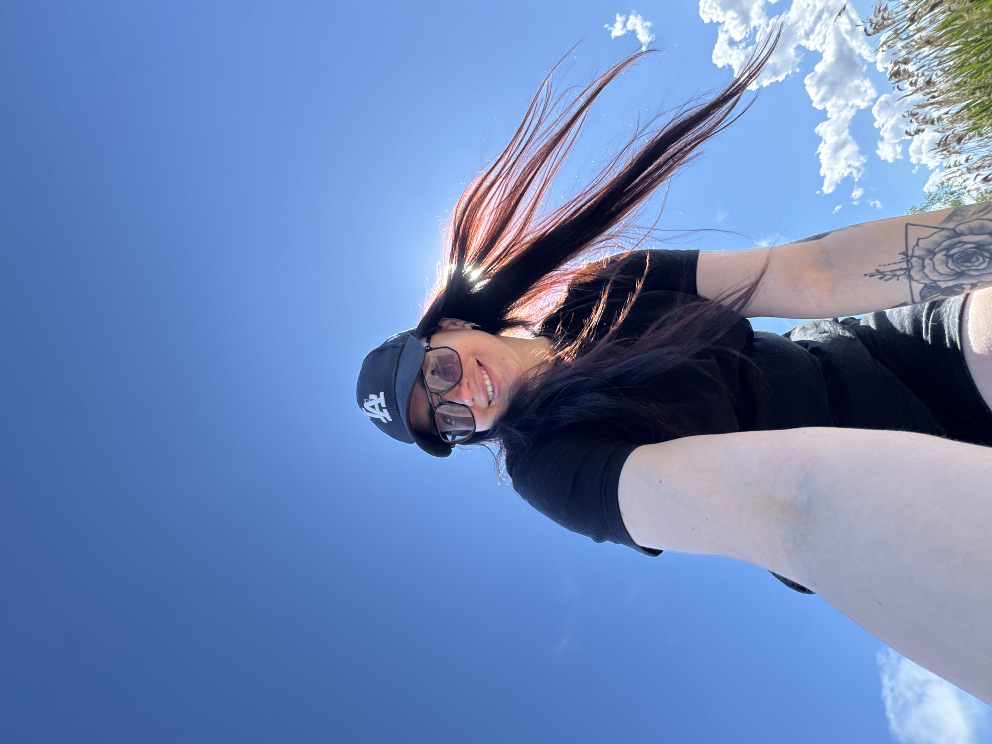 A low-angle medium shot depicts a woman with shoulder-length, dark-brown hair with red highlights. She is wearing a black t-shirt, a black Los Angeles Dodgers baseball cap, and dark-rimmed glasses. Her hair is partially obscuring her face and the sun. Her right arm is extended toward the camera, displaying a tattoo on her forearm featuring a rose and geometric shapes. In the background, there is a clear blue sky with some white clouds and a small patch of green foliage in the upper right corner. Location: Unknown, Unknown.