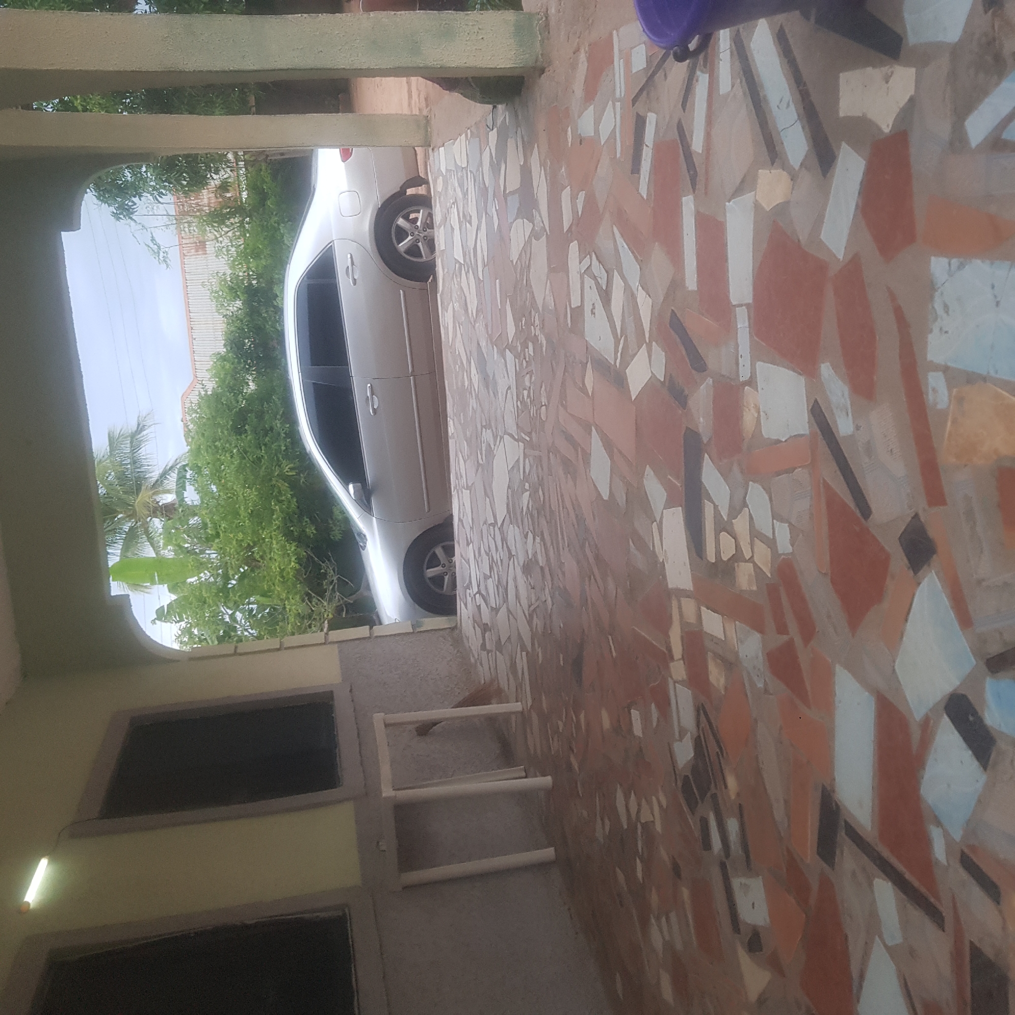The image shows an outdoor scene, possibly a covered porch or walkway, with a light-colored car parked in the background. The ground is paved with a mosaic of irregularly shaped tiles in shades of brown, beige, white, and blue. To the left, there is a light yellow wall with two dark rectangular openings, possibly windows or doors. A fluorescent light tube hangs on the left side of the wall, emitting a soft glow. In the background, there are trees and foliage, and the sky is visible, suggesting it is daytime. The overall impression is a snapshot of a residential or semi-public outdoor area. There are no people visible in the image.