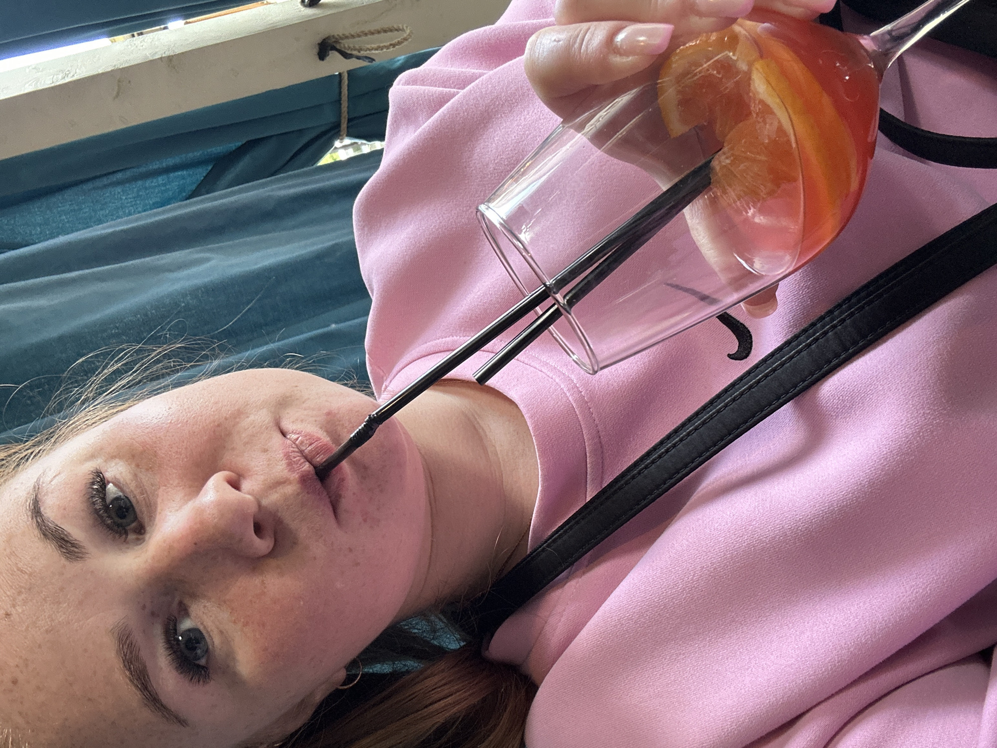 A Caucasian female with fair skin and freckles is shown in a close-up, angled selfie, holding a stem glass containing a red-orange beverage with citrus slices and two black straws. The woman has blue eyes, dark eyebrows, and red hair tied back. She is wearing a light pink sweater with a black strap crossing her chest, presumably a bag strap. The woman has one straw in her mouth and is drinking from the glass. The background features a teal fabric, possibly curtains, with a white wooden beam above. The location is not explicitly stated as Chaikovsky, Russia and is not discernible from the image.