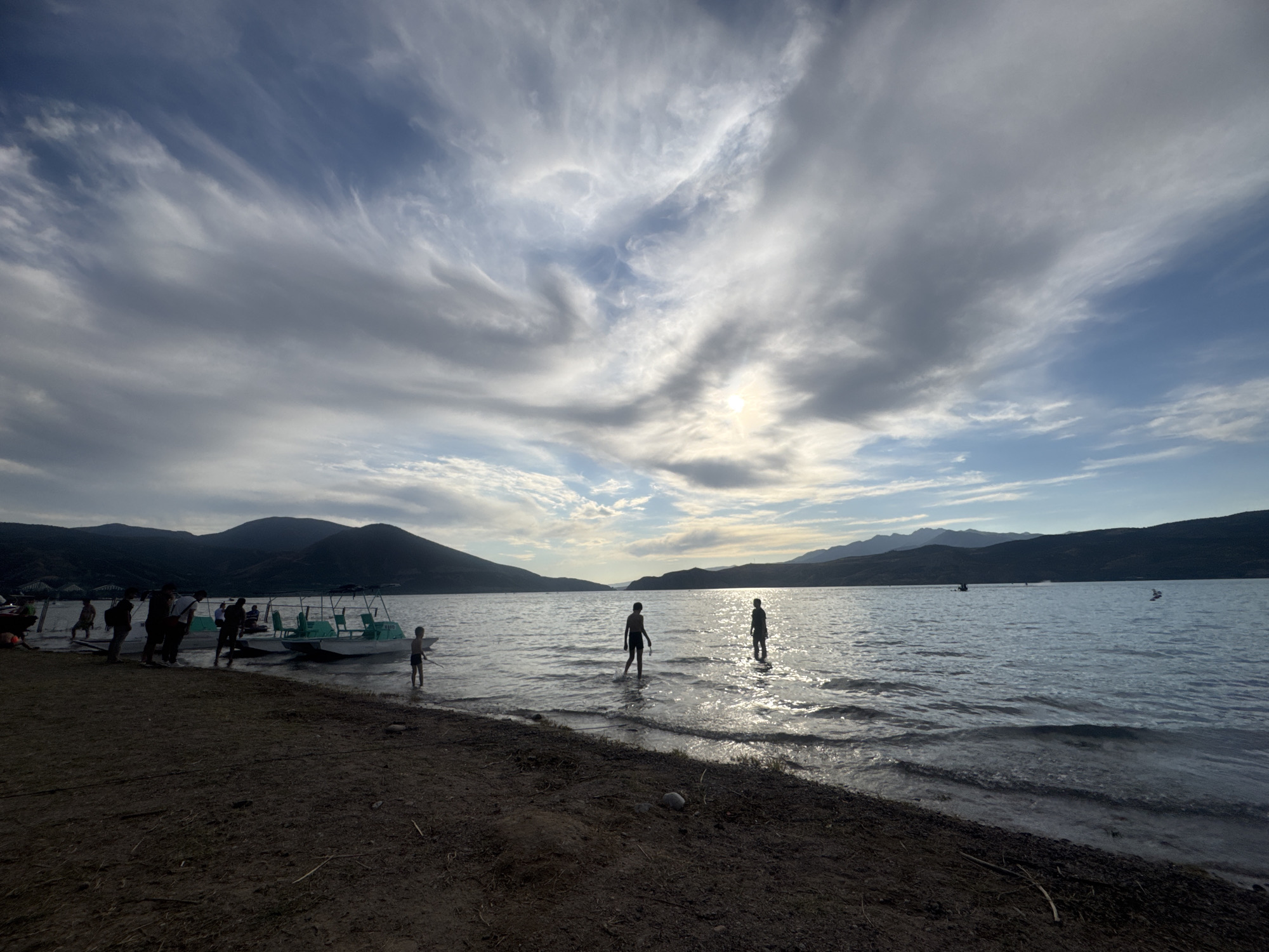 A wide shot captures a silhouetted scene at a lake's edge under a dynamic sky. Dark, undulating hills form the backdrop, with a vast expanse of water stretching towards them. The water reflects the bright, partially obscured sun, which is breaking through a dramatic sky filled with wispy, textured clouds in shades of white, gray, and blue.
In the foreground, a dark, sandy shoreline is visible. Several boats, likely for rent or personal use, are docked or partially in the water along the left side of the frame. A group of people, mostly in silhouette, are gathered near the boats and on the shoreline. Some appear to be boarding or disembarking from the watercraft.
Further out in the lake, three figures, also in silhouette, are standing in the shallow water. Two are children, and the third, taller figure, is an adult. They are positioned towards the right of the frame, with the sun's glare creating a luminous halo around them. Ripples and gentle waves lap at the shore and surround the figures in the water. The overall atmosphere is one of natural beauty and recreational activity.