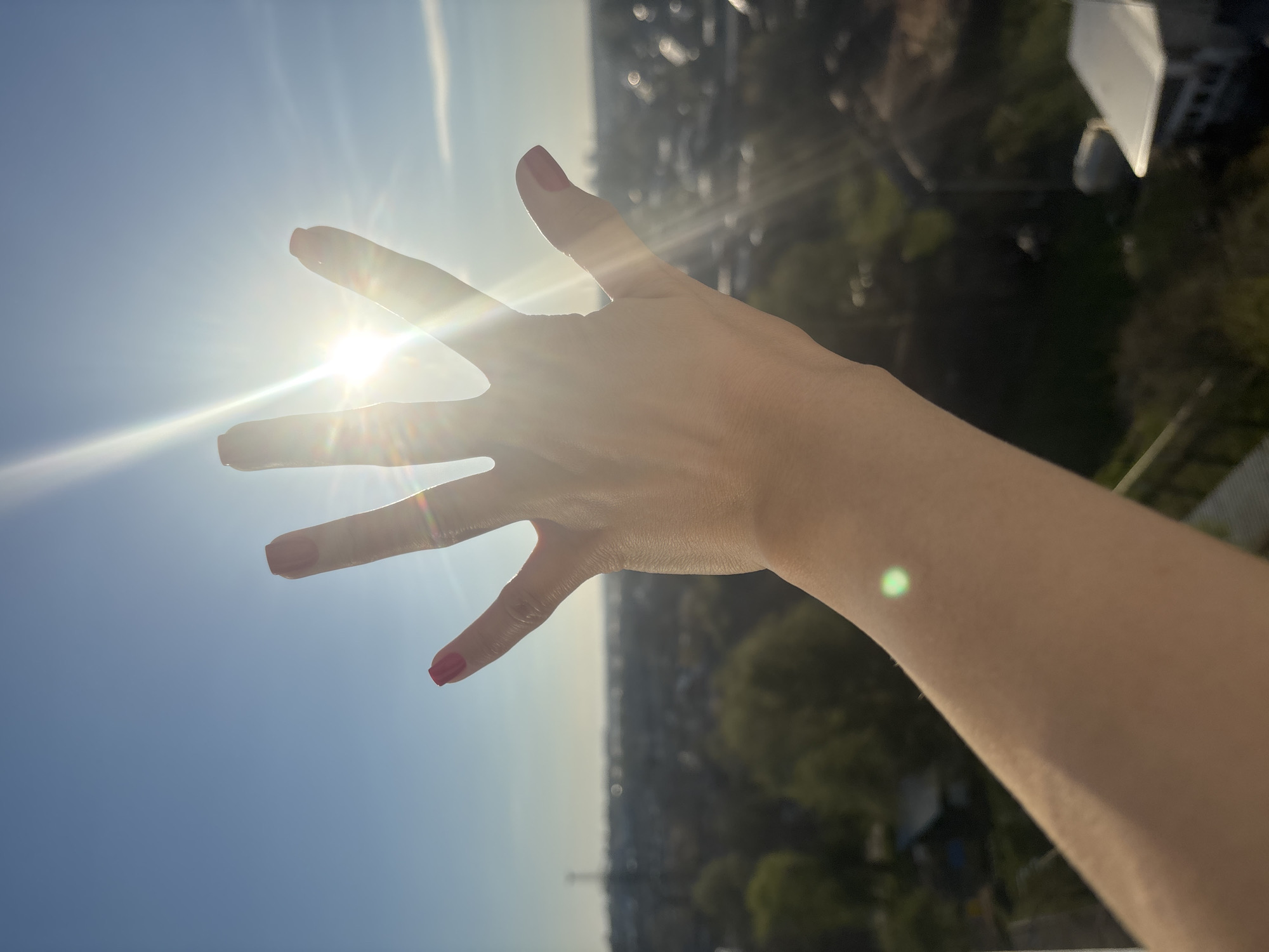 A woman's hand with red nail polish is outstretched toward the sun in Chaikovsky, Russia. The sun is partially obscured by the hand, causing bright light rays to emanate outward. The background is a cityscape with blurred buildings and trees under a clear blue sky.