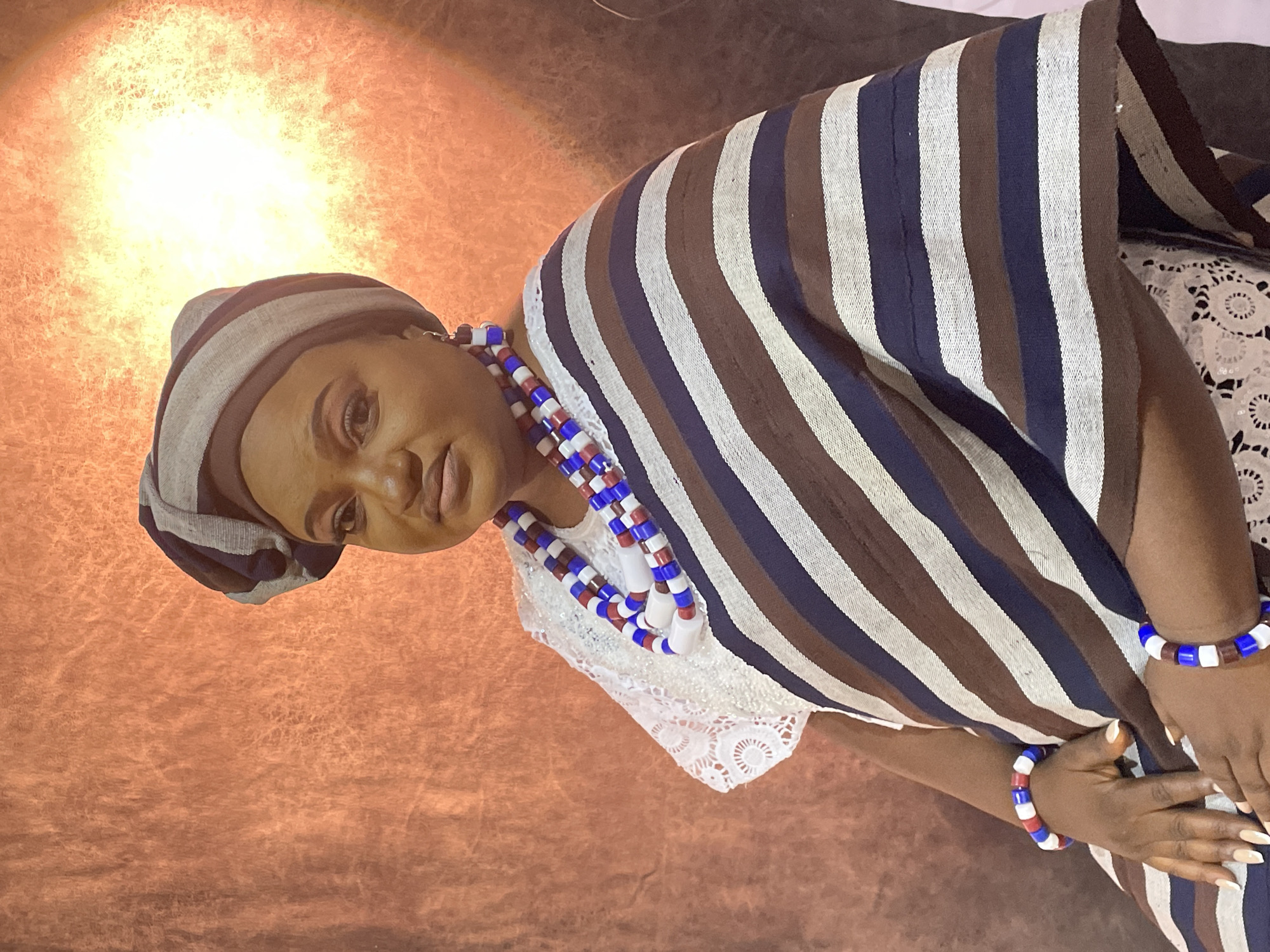 The image shows a woman indoors wearing traditional attire. She is wearing a head covering and a striped shawl or garment. She also has beaded necklaces and bracelets with red, white, and blue beads. The background appears to be a textured wall, possibly lit with warm light. There is no information to determine if it is in Barakin Akawo, Nigeria.