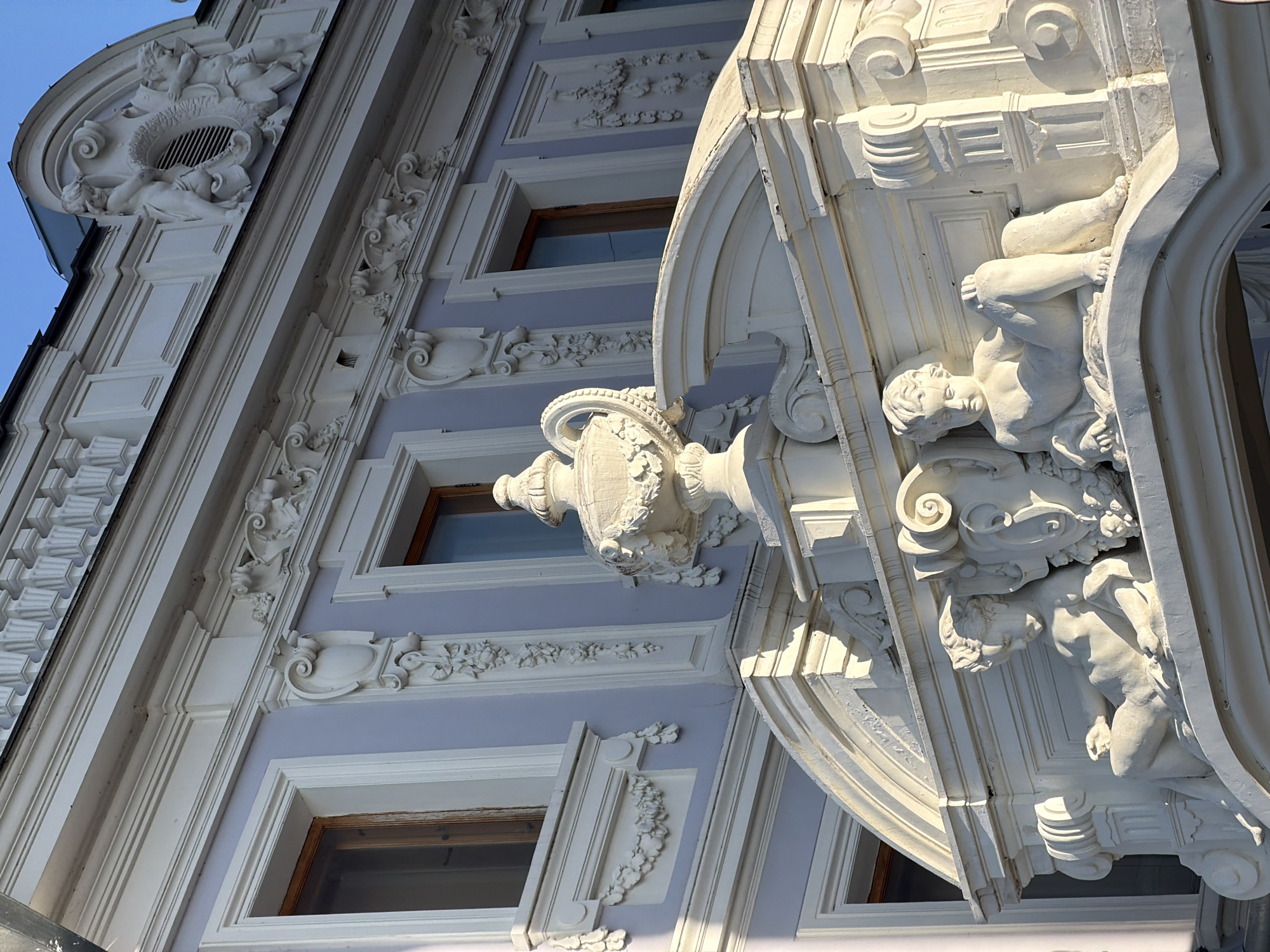 The image shows a close-up of the facade of an ornate building in Nizhny Novgorod, Russia. The building features classical architectural elements, including decorative moldings, sculpted figures, and an urn-shaped ornament. Two cherubic statues are prominently displayed on a curved ledge, resting on a white, sculpted surface. The building's facade is painted a pale lilac with white decorative trim around the windows and architectural features. The sky is visible in the upper right portion of the image, a clear blue. The overall impression is one of historical grandeur and detailed craftsmanship.