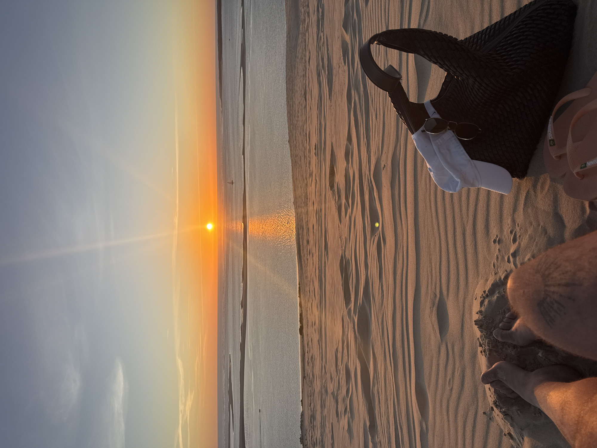 A first-person perspective shot captures a sunset over the Dune of Pilat in La Teste-de-Buch, France. The foreground is dominated by rippled sand dunes. In the mid-ground, a black woven bag is placed on the sand, with a white cap and sunglasses hanging from its strap. Two feet, one with a tattoo, are visible at the bottom left of the frame, partially buried in the sand. A pair of orange flip-flops rests in front of the bag. The background features a wide expanse of water, with the sun setting on the horizon, casting a warm orange glow across the sky and reflecting on the water's surface. The sky transitions from a pale blue at the top to a warm orange near the horizon, with thin wisps of clouds. A distant sailboat and a boat are visible on the water.