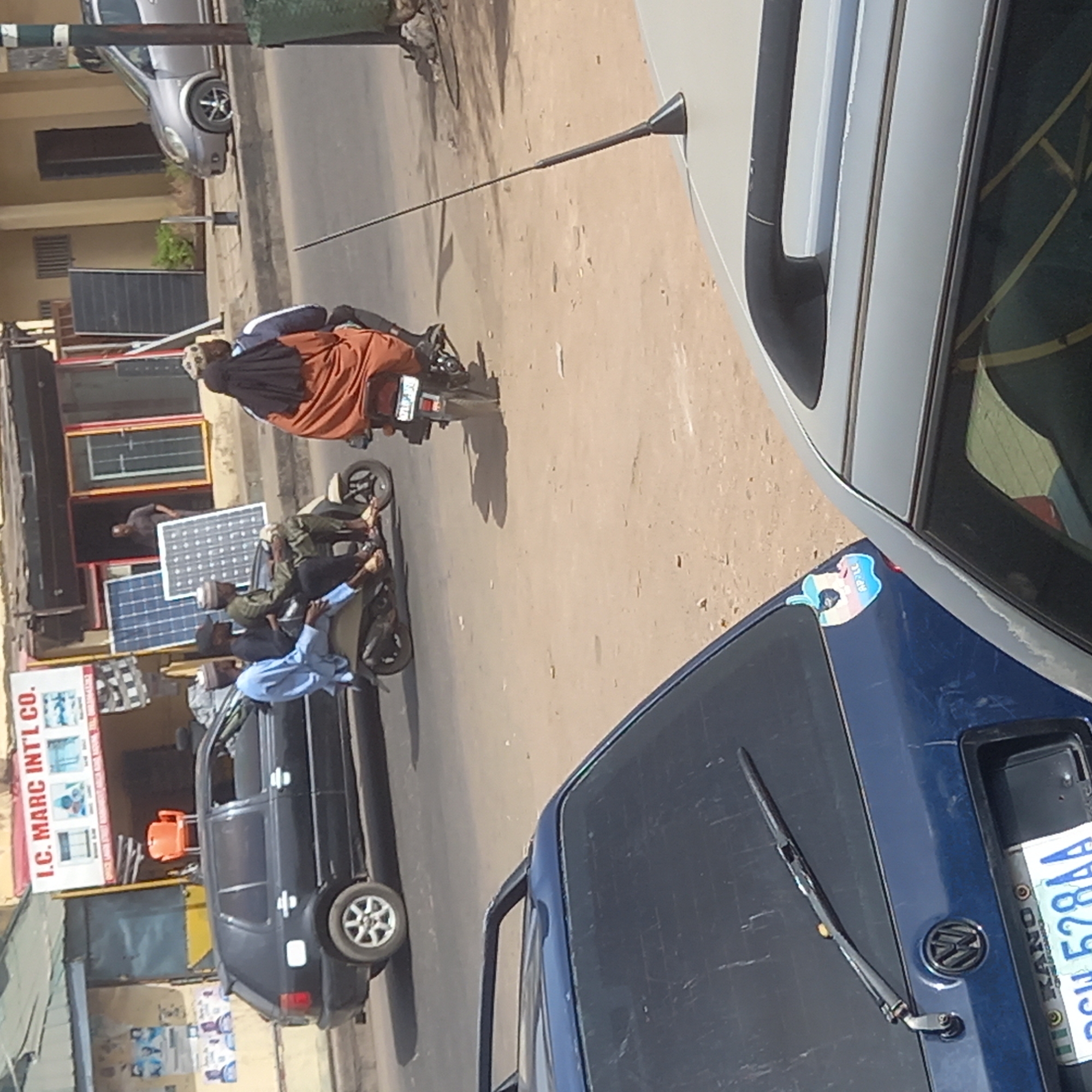This tilted image captures a vibrant street scene in what appears to be Sardauna, Nigeria, under bright daylight. The perspective is from a slight elevation, looking down at a street with several vehicles and people.
In the immediate foreground on the right, a dark blue car's hood and windshield are visible. A distinctive sticker featuring an apple logo with the transgender flag colors and the word "APPLE" is affixed to its hood. A Nigerian license plate with "KANO" and "GH 528AA" is also present on this vehicle. Extending from the top right, a grey sedan is partially seen, hinting at other parked or moving vehicles.
The road itself is a light, dusty brown surface, with strong shadows cast by people and vehicles, suggesting clear, sunny weather, likely mid-morning or afternoon. Several people are engaged in daily activities. On the left side of the street, a dark grey hatchback is parked near commercial establishments.
Central to the image are multiple people on motorcycles. One motorcycle carries three individuals: the driver in a green top and cap, and two passengers, one of whom wears a light blue shirt. They appear to be stopped or moving slowly in traffic. Further down the road, another person on a motorcycle, dressed in an orange robe over a dark top and a head covering, is also visible.
Along the left side of the street, storefronts are visible. One shop prominently displays multiple large solar panels, indicating it deals in solar energy products. Above this shop, a sign clearly reads "I.C. MARC INT'L CO." A person can be seen standing in the doorway of one of these shops, observing the street. The scene depicts a bustling, everyday street environment characteristic of the region.