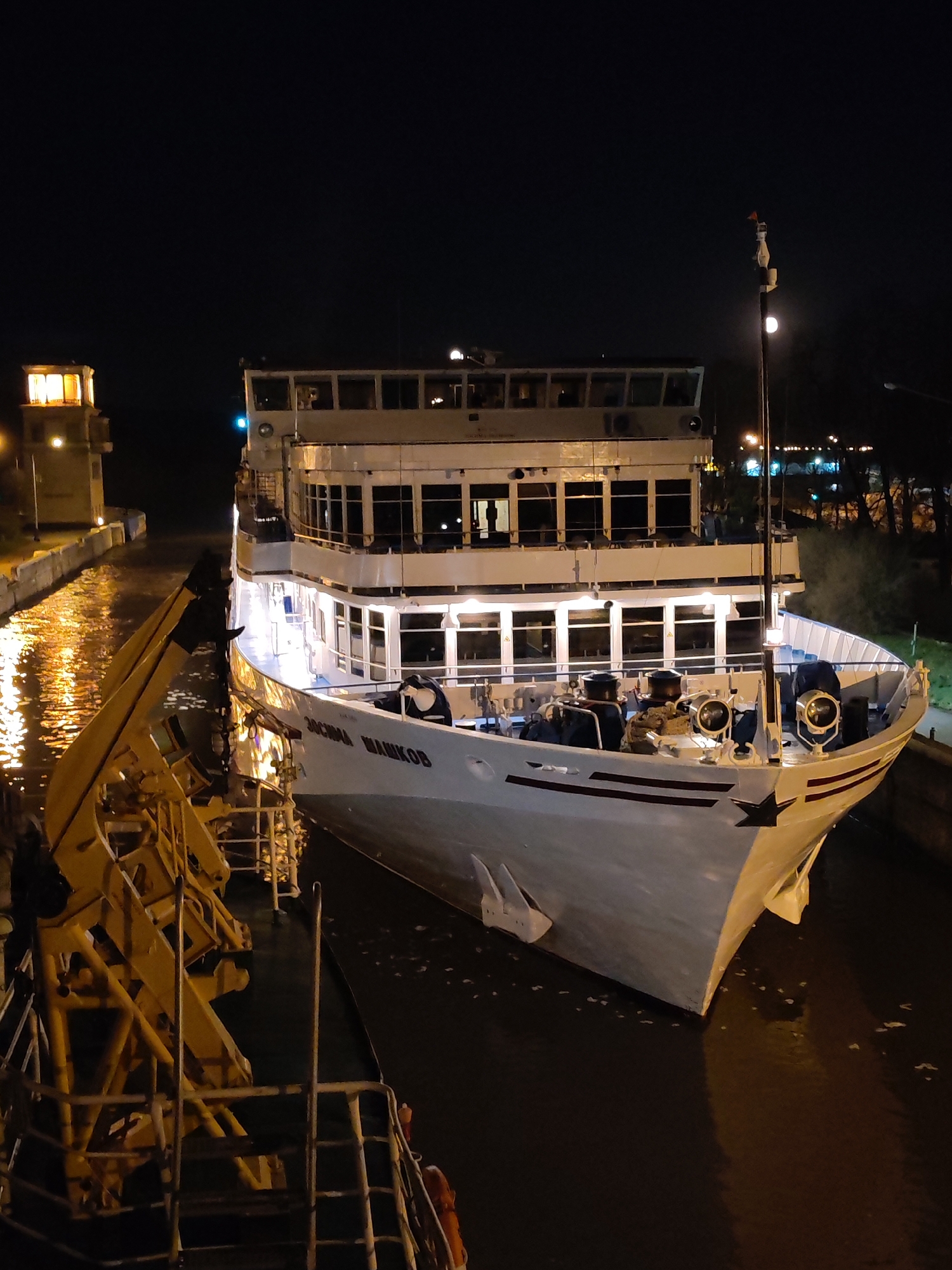 At night, the "ZOSIMA SHASHKOV" ship is positioned in the lock of the Moscow Canal in the city of Yakhroma, Russia. The multi-decked, white ship with lighted windows sits low in the water. A yellow metal structure is alongside the ship on the left. A light tower is visible on the left. Trees and streetlights are on the right. The water reflects light.