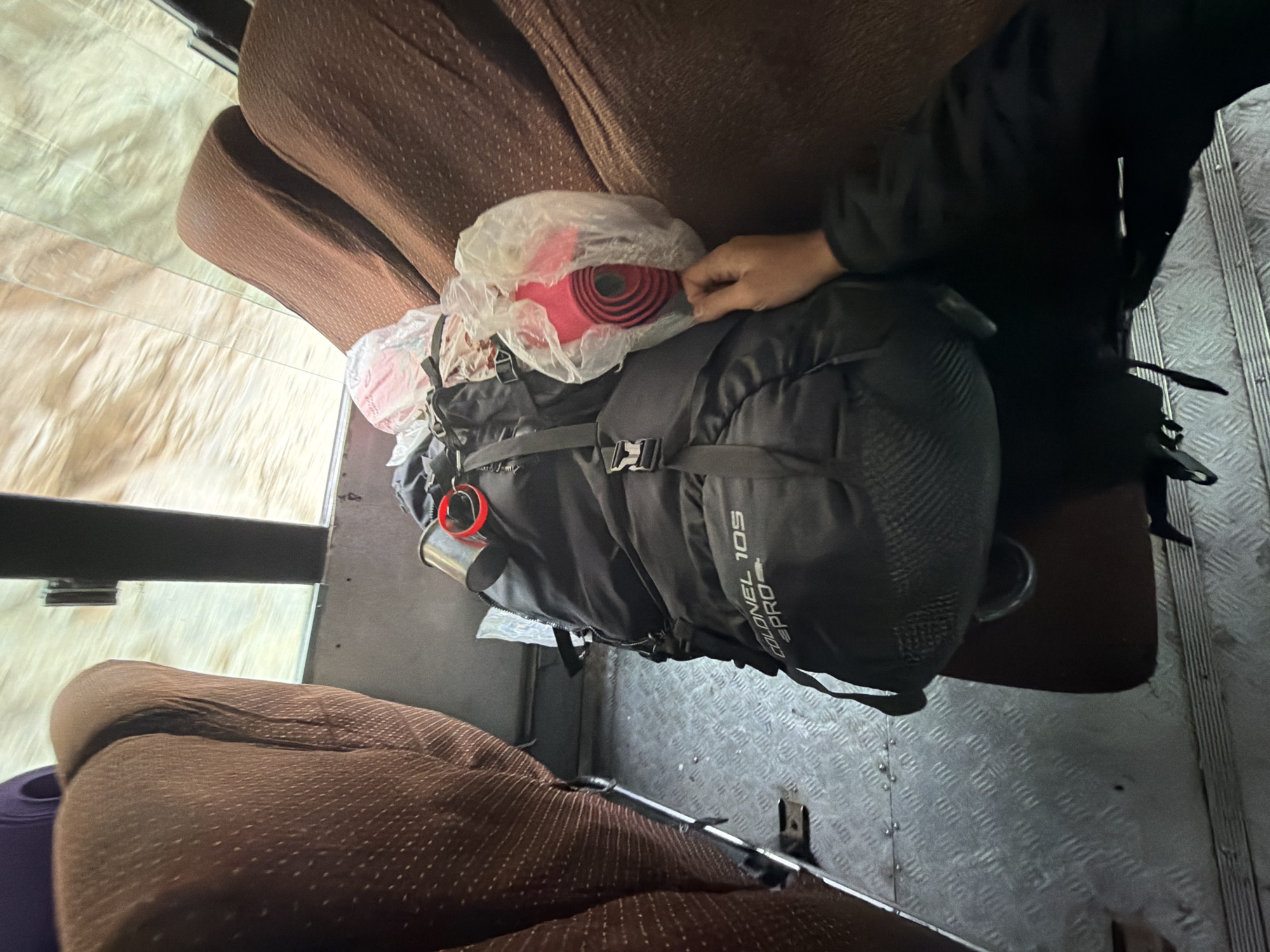 A black backpack with "COLONEL 10S PRO" printed on it sits on the floor of a bus, between two brown seats. The backpack has various straps and buckles. A metal water bottle is clipped to the top left of the backpack. To the right of the backpack, a person's hand is visible, reaching into a clear plastic bag containing a rolled-up red object. The floor of the bus is made of diamond-patterned metal. The seats appear to be upholstered in brown fabric with a textured pattern. Outside the bus window, a blurred image of what appears to be a natural landscape or body of water is visible. The scene suggests a journey or travel. The location is identified as the city of Gayk, India.