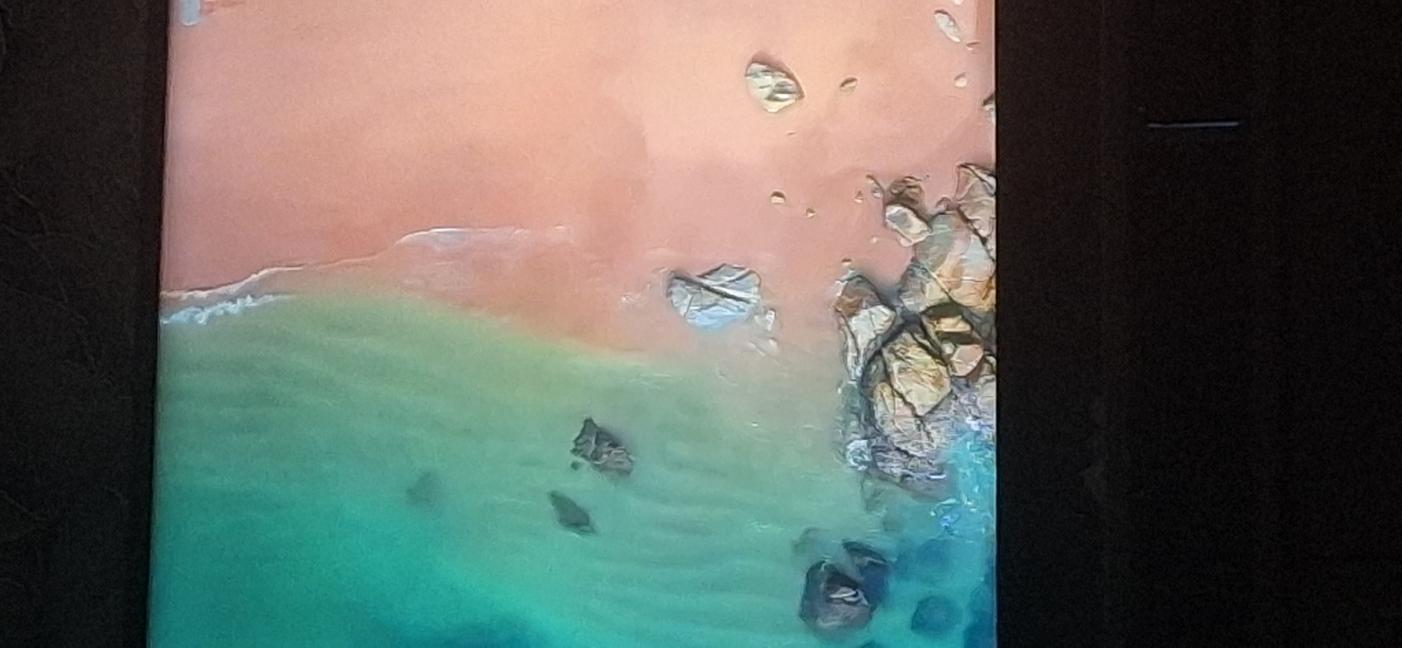 The image is an aerial view of a coastline. The water is a vibrant turquoise and appears clear, with some dark rocks visible beneath the surface. The sand is a pale pinkish-orange and is wet near the waterline, indicating the tide is coming in or going out. Several scattered rocks and a cluster of larger rocks are present on the shore. The top and bottom portions of the image are black, suggesting the photograph was taken from a device with a dark frame or bezel. There are no people or man-made structures visible in the scene. The location in the city of Bila Tserkva, Ukraine cannot be determined from this image.