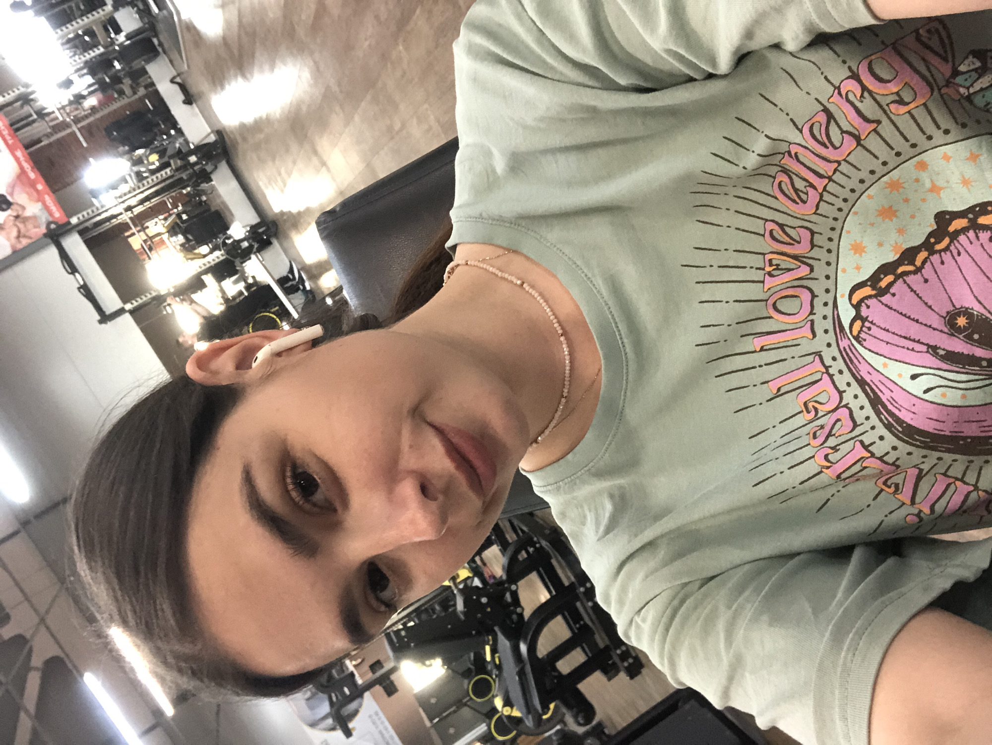 The image is a selfie taken by a young woman inside a gym. She is wearing a light green t-shirt with a graphic print on it. She has her hair pulled back and is wearing white earbuds. The background shows gym equipment, including weight machines and a squat rack. The flooring appears to be made of wood. There are fluorescent lights on the ceiling, and the overall lighting suggests it is daytime or a well-lit indoor space. The woman is looking at the camera and smiling slightly. The location is identified as the city of Chaikovsky, Russia.