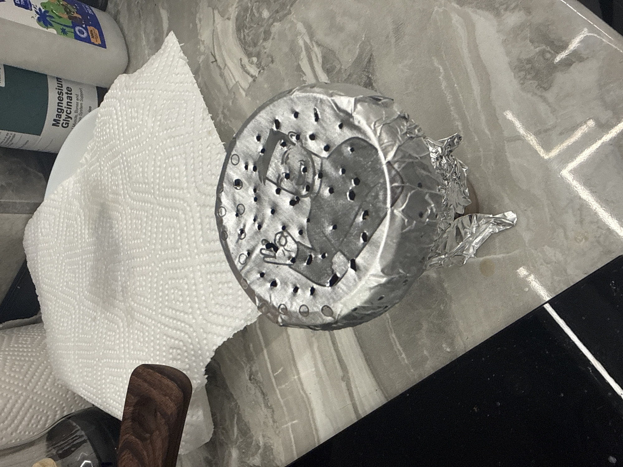 The image shows a collection of items on a patterned surface, likely a kitchen countertop.
**Objects:**
* A white bottle labeled "Magnesium Glycinate" is partially visible in the upper left corner.
* A wadded white paper towel occupies the left side of the image.
* A cylindrical metal object, possibly a hookah bowl, is centrally located. The object is covered with foil, which has a design of a person and has small holes punched into its surface.
* A dark brown wooden object, possibly a handle, appears in the lower left corner.
* A black rectangular object is visible in the lower right corner.
**Environment:**
* The surface the objects are on is a light gray with a marble-like pattern.
The image provides no information about the location being Burgas, Bulgaria.