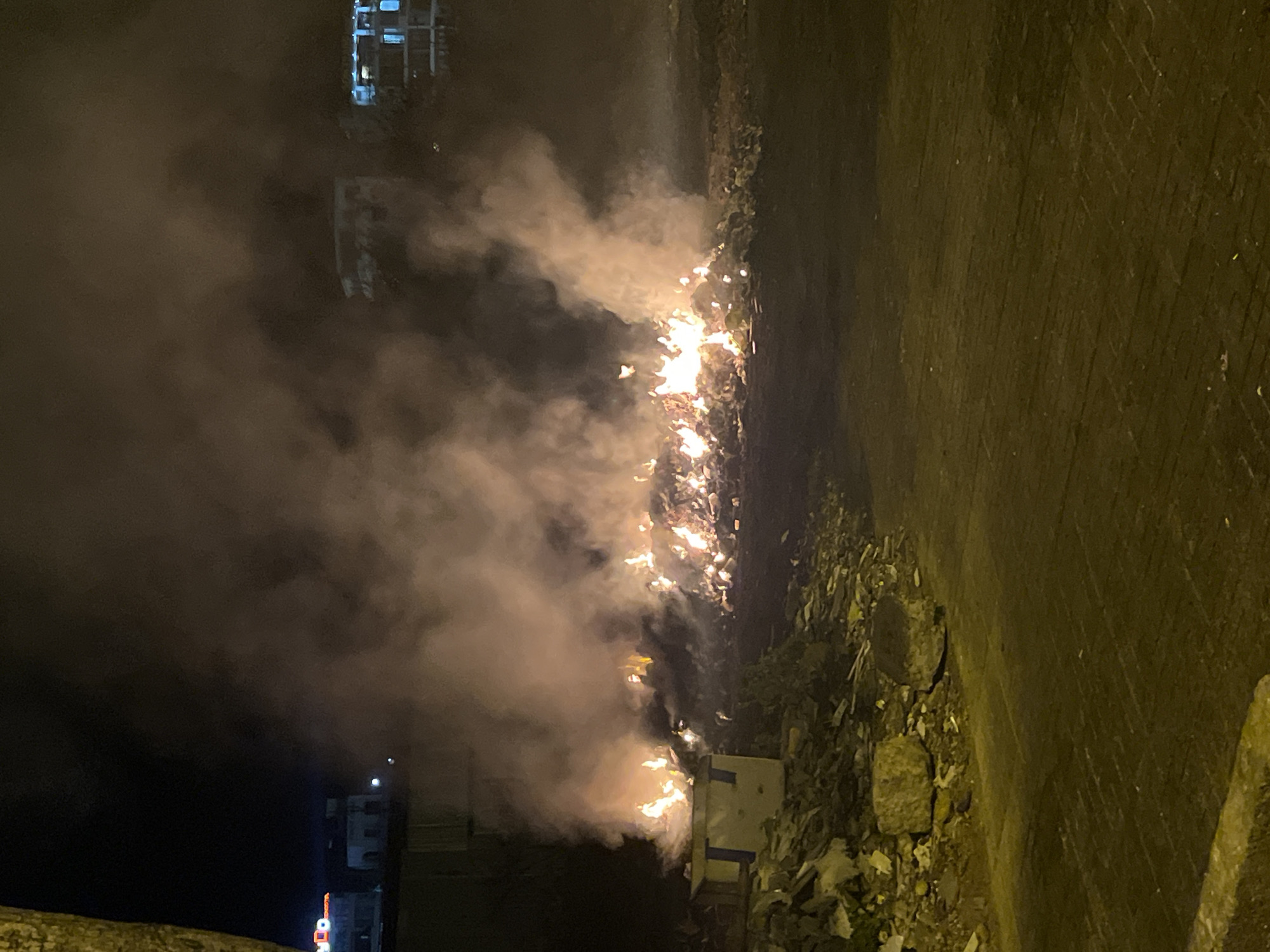 At night in Bac Nha Trang Ward, Vietnam, a fire burns along the side of a street. The flames and smoke are obscuring buildings in the background. To the right, a wall follows the length of the image, which looks to be adjacent to a street with debris or waste. In the immediate foreground, some shrubbery or debris is visible.
