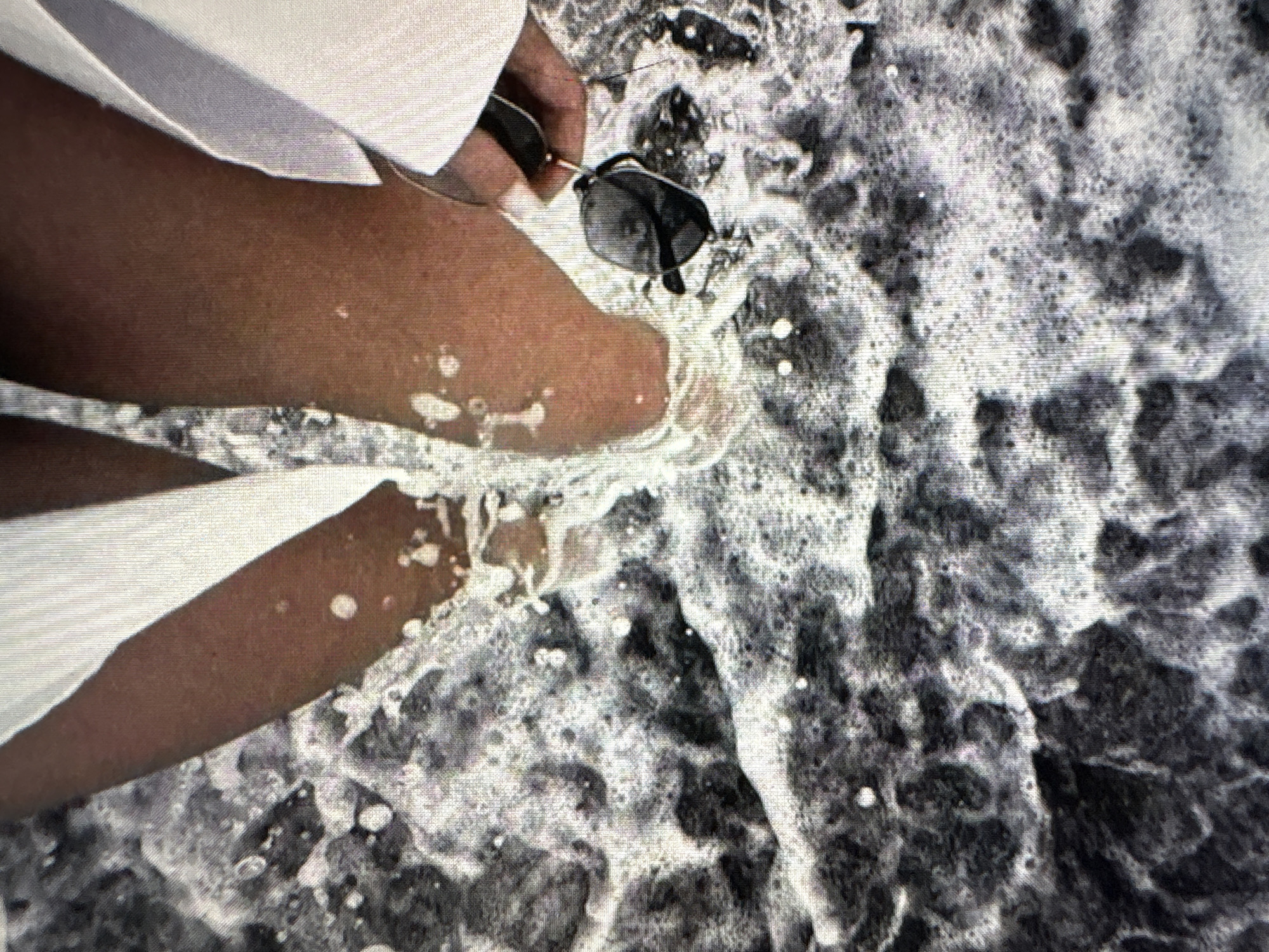 A person's legs are partially submerged in foamy ocean water. They are wearing white clothing and holding a pair of sunglasses. The water is dark and frothy with white foam and small bubbles. The image is taken from a high angle looking down at the person's legs.