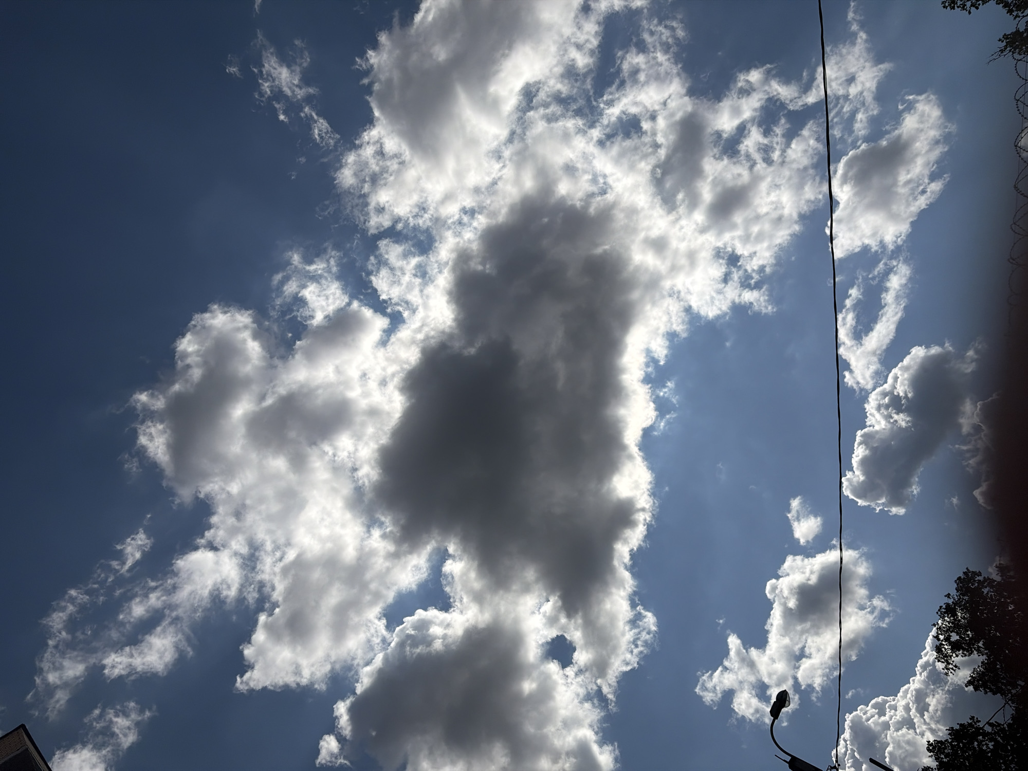 The image shows a sky filled with a mix of dark and white clouds against a blue sky. The sun is not directly visible but is strongly implied by the illumination and shadows on the clouds. In the foreground, the upper branches of dark green trees are visible at the bottom of the frame, along with a barbed wire fence just above them. To the left, a lamppost with a curved arm is partially visible, and a dark, thick wire runs horizontally across the middle of the frame. The scene appears to be outdoors in Sverdlovskiy, Russia, likely in a semi-urban or suburban area given the presence of trees, a lamppost, and a fence, under a dynamic sky.
