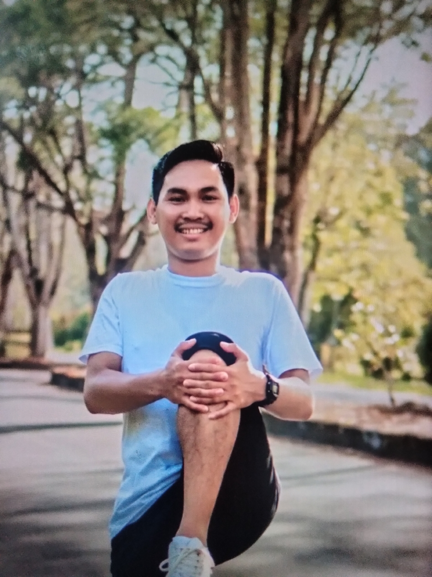 A young man in a light blue t-shirt and black shorts is stretching his left leg outdoors. He is holding his knee with both hands, with his right leg bent and his foot resting on the ground. He is smiling and looking at the camera. The background is blurred, showing trees and a paved path. The location is described as Payagaboh, Indonesia.