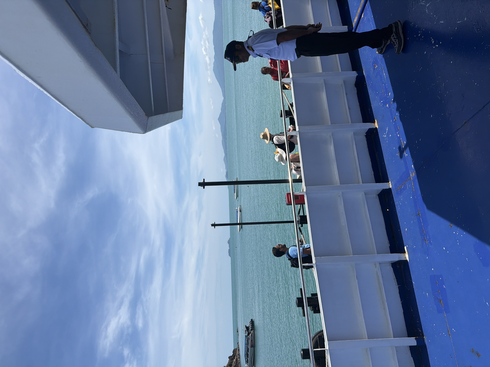 On the deck of a ferry, a man in a uniform stands near the railing overlooking the ocean. Other passengers are visible behind him. The ocean stretches to the horizon, with islands in the distance. The sky is blue with scattered clouds. The ferry's structure is visible, including white railings and a blue floor. The location is likely near Baan Tai, Thailand, given the tropical environment and ocean view.
