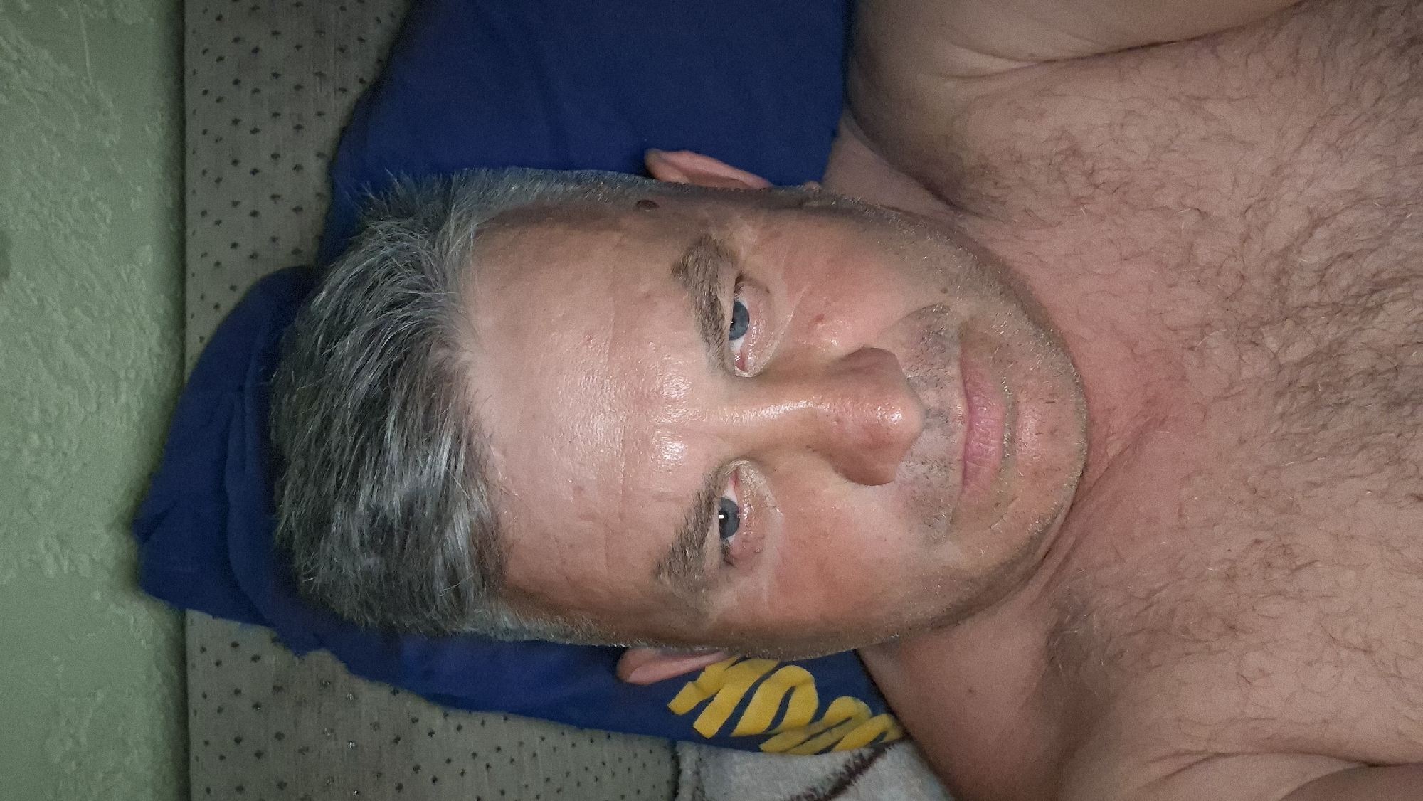 A close-up, overhead selfie of a shirtless man with graying hair and a fair complexion is depicted. The man has blue eyes, stubble, and is looking directly at the camera with a neutral expression. His skin appears flushed and slightly oily. He is lying down, with a dark blue pillow visible behind his head and a patterned fabric that may be part of bedding or furniture. A portion of a yellow and blue patterned object, possibly a towel or fabric, is visible to the left of the man's face. The background appears to be a wall with a textured green finish. The overall lighting suggests an indoor setting, possibly at night. The image was taken in the city of Voinova, Moldova.