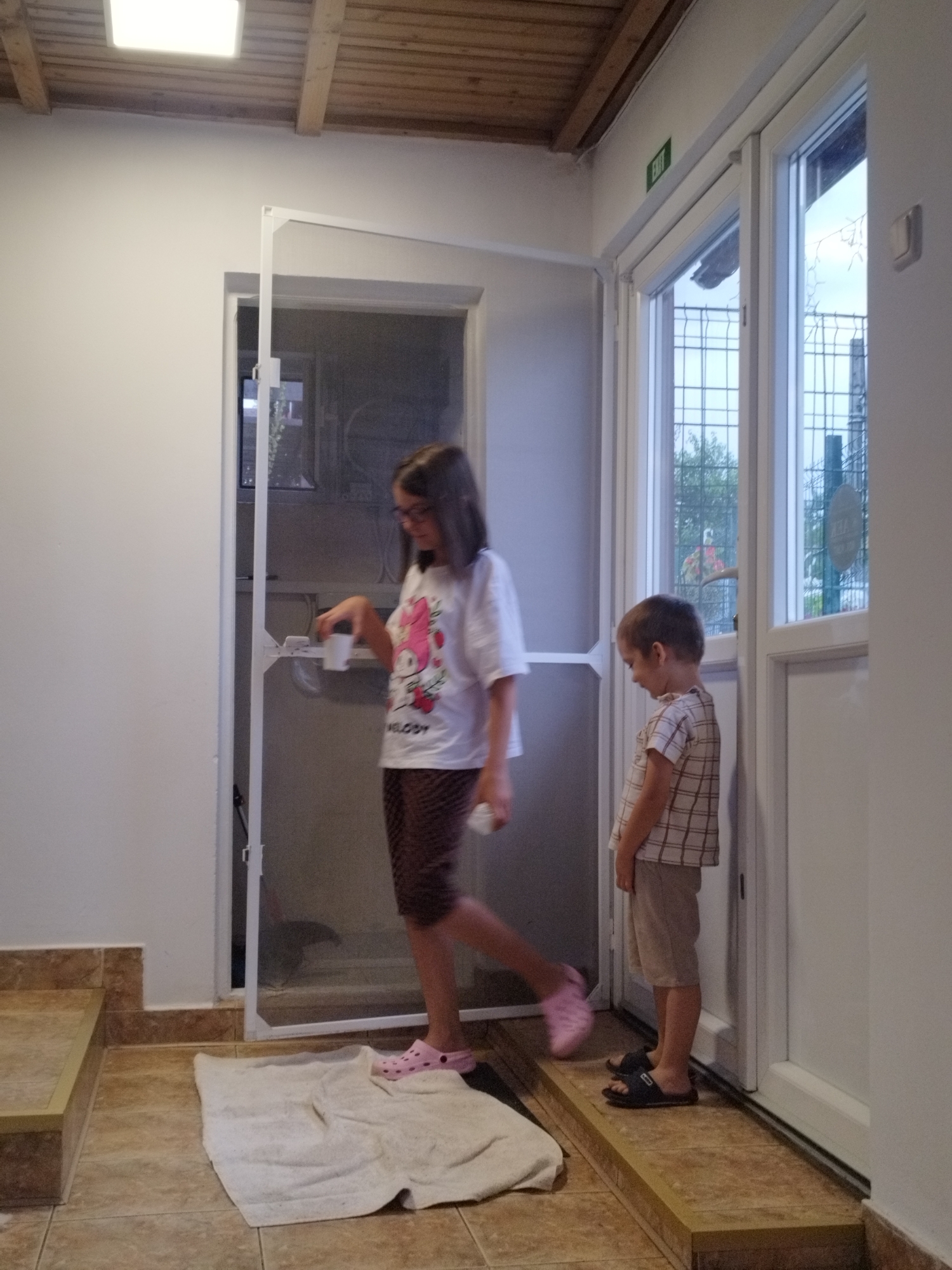 In Bucharest, Romania, a young girl with dark hair and glasses, wearing a white t-shirt with a pink character and brown patterned leggings, stands to the left of a screen door. She is holding a white cup in her right hand. To her right, a younger boy with light brown hair, wearing a plaid shirt and beige shorts, stands facing away from the viewer, looking down. He is wearing dark sandals. A light-colored towel is placed on the tiled floor in front of the screen door. The environment appears to be an indoor space with white walls, a wooden ceiling with exposed beams, and large windows. An "EXIT" sign is visible above the windows. The floor is tiled in shades of brown and beige.