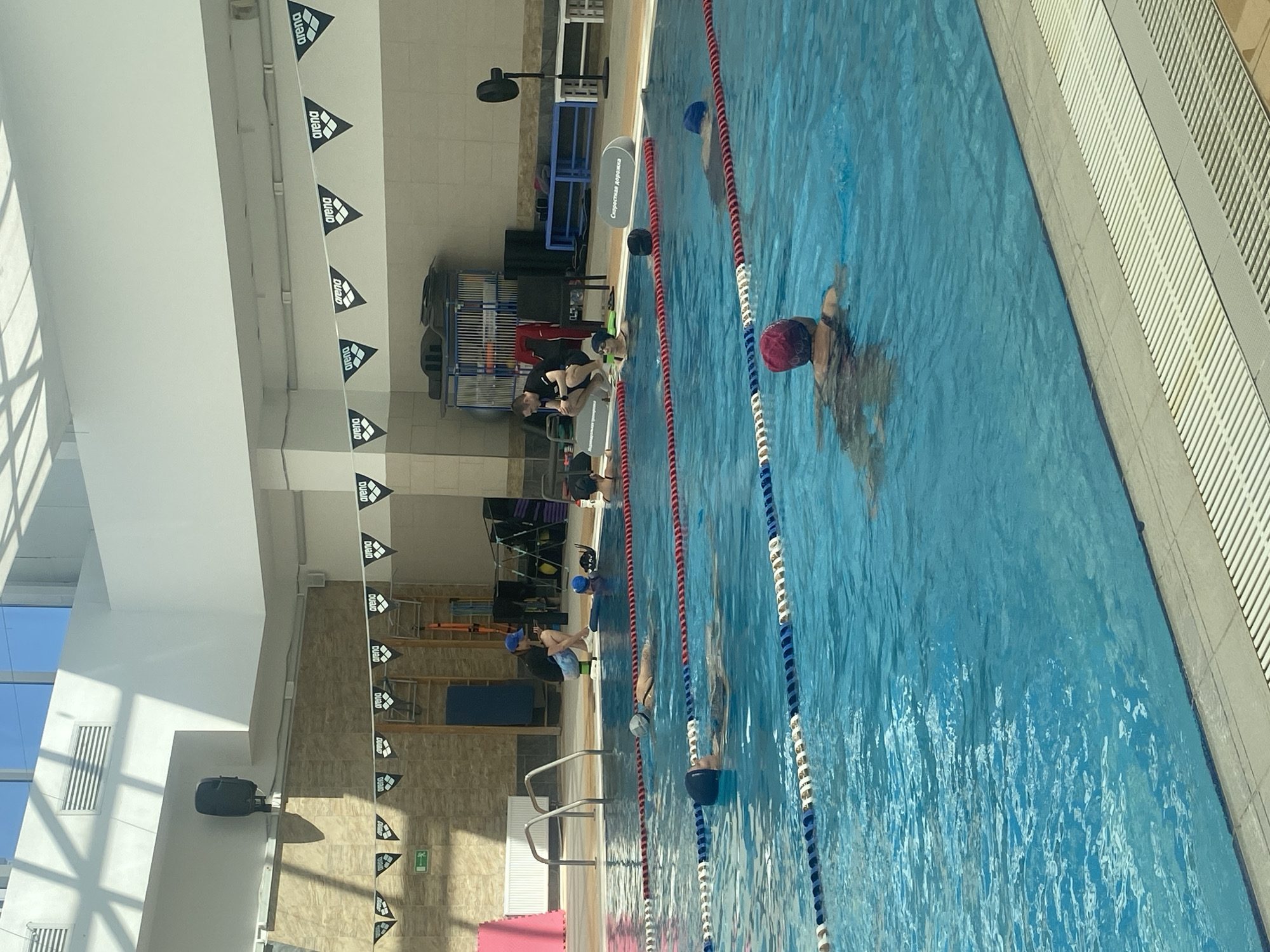 The image shows an indoor swimming pool. Several people are either in the pool swimming or standing alongside it. The pool is divided into lanes by ropes. The pool is located in Yekaterinburg, Russia. Banners with the word "arena" hang overhead. Exercise equipment can be seen near the edge of the pool. The pool has tiled walls and a grated walkway.