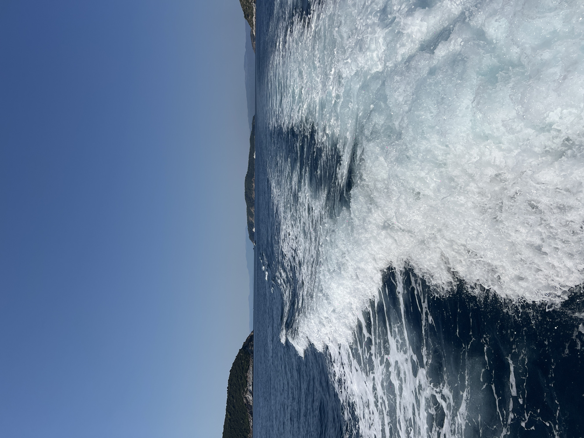 The image captures a view from a boat moving through the Aegean Sea, leaving a prominent wake. The water is a deep blue, with the wake creating frothy white patterns extending behind the vessel. In the distance, green, tree-covered islands are visible on the horizon, under a clear, bright blue sky. One of these islands, likely Skiathos, Greece, is characterized by its lush vegetation and a coastline that slopes down to the sea. The overall scene suggests a sunny day at sea, likely near the island of Skiathos, given the geographical features depicted. There are no people or specific landmarks identifiable in the image beyond the natural environment.