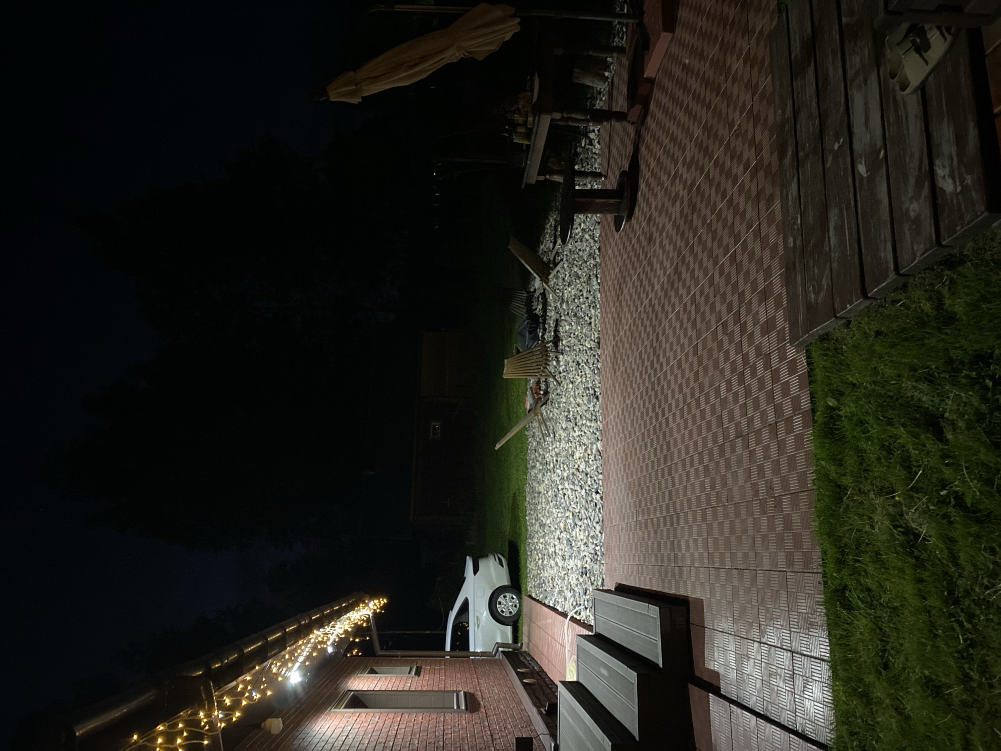 The image depicts an outdoor residential area at night in Копнино, Russia.
On the left, a two-story brick building with two visible windows and dark window frames is illuminated by warm yellow string lights draped along its roofline and by additional downward-facing lights. A white sedan is parked on a strip of grass adjacent to the house, partially visible. Dark wooden steps descend from the house to a patio.
The patio features reddish-brown interlocking tiles with a patterned surface. Beyond the patio, a large area is covered with light-colored irregular stones/gravel. Two wooden slatted lounge chairs are positioned on this gravel, facing a dark circular object, possibly a fire pit, between them.
On the right side of the patio, a rustic wooden table holds several dark-colored bottles and other items. Adjacent to it, a large, beige, folded patio umbrella stands upright in a base. A small, dark, round table with a central pedestal is situated on the patio in front of the larger table. In the immediate foreground, to the bottom right, dark wooden planks are laid on the grass, with a pair of light-colored open-toed slippers resting on them.
The background consists of dark trees and a dark, possibly wooden, fence structure. The sky is dark and cloudless. Green grass is visible in the foreground and bordering the gravel area. No individuals are visible in the scene.