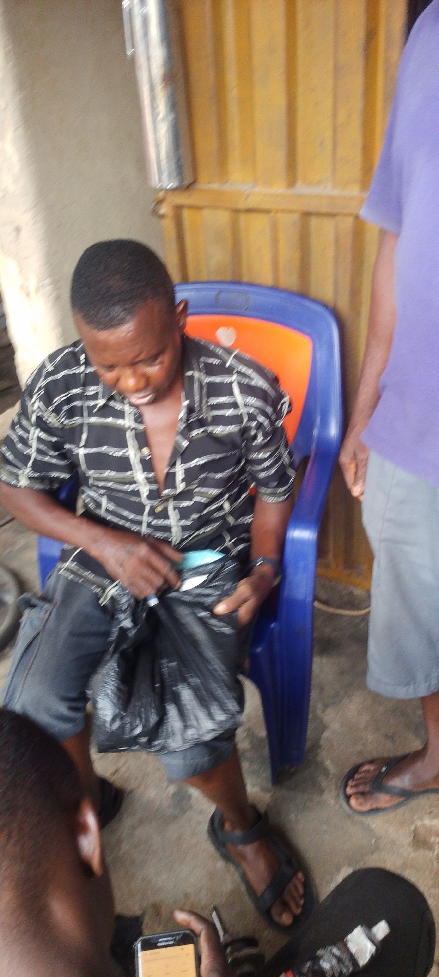 In Lokoja, Nigeria, the image depicts a man seated in a blue and orange plastic chair. He is wearing a short-sleeved black and white patterned shirt and dark jeans cropped at the calves. He is holding open a black plastic bag, appearing to retrieve something from it.
Part of a man wearing a purple shirt and gray shorts is visible on the right, with his feet in sandals. At the bottom, parts of two other individuals are visible, one with a cell phone in hand. The environment appears to be an indoor or covered space with a corrugated metal wall in the background. A silver, cylindrical object is affixed to the wall. The floor is bare and dusty.