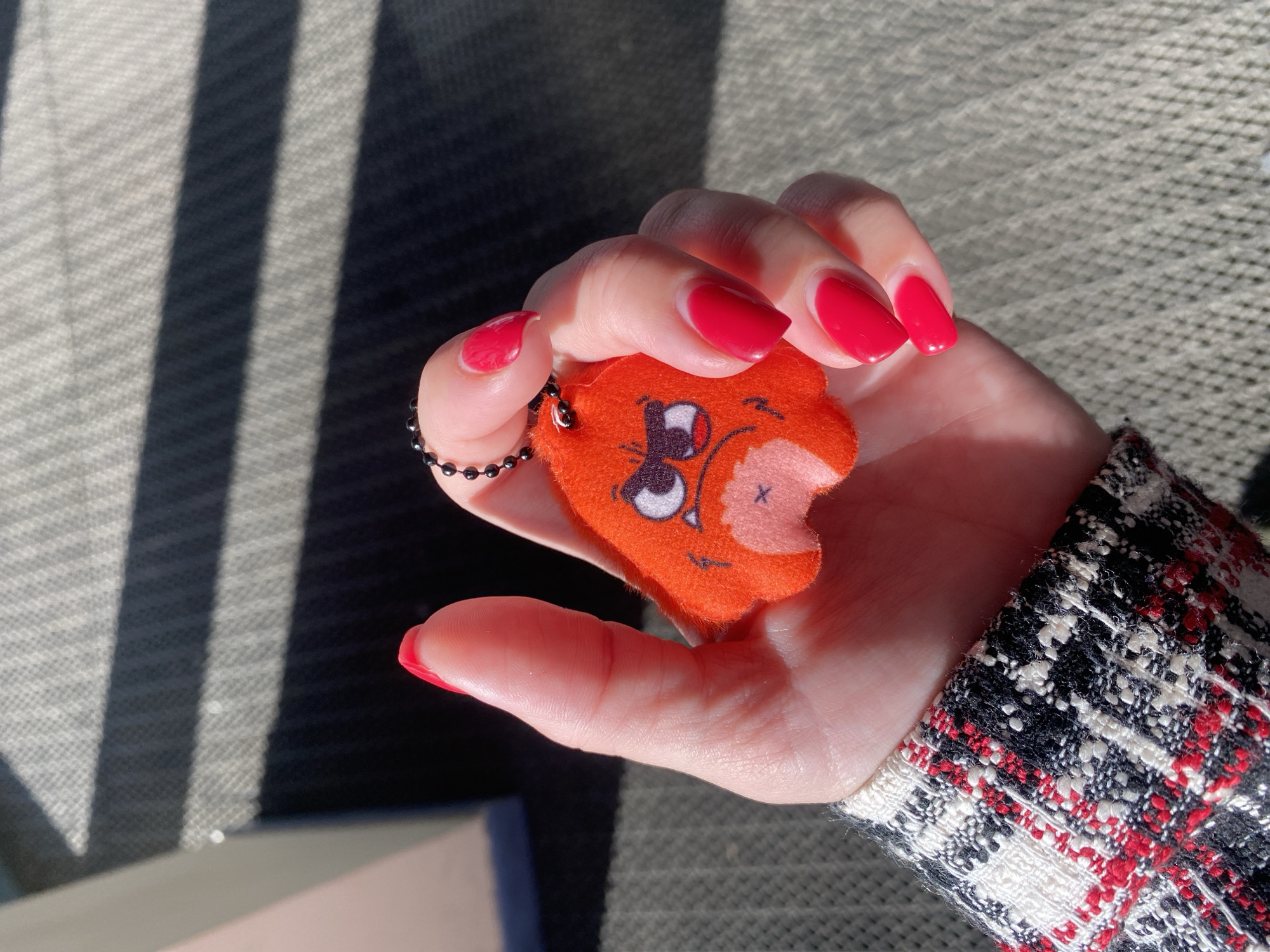 A hand with red manicured nails is holding an orange plush keychain in the shape of a character with a bite taken out of its ear. The keychain is attached to a black ball chain. The hand is positioned in front of a surface with alternating light and dark stripes and is wearing a black, white, and red patterned jacket. The location is Izhevsk, Russia.