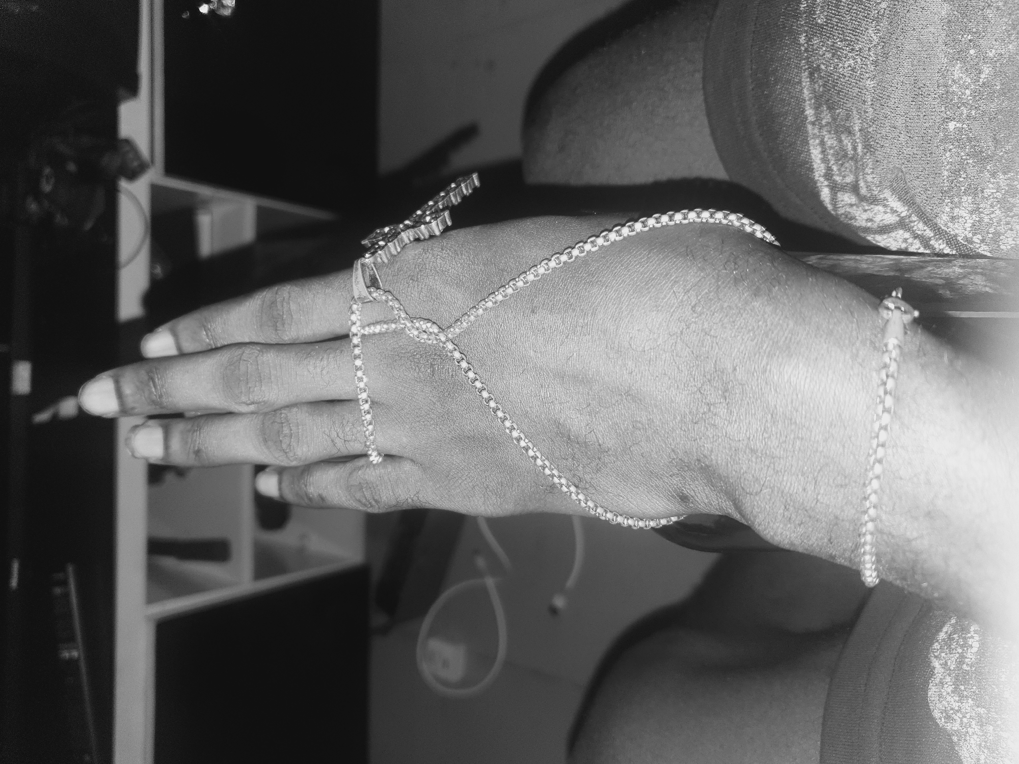 A close-up, grayscale image shows a hand adorned with a decorative hand chain. The hand, which appears to belong to an adult, is positioned with the fingers extended towards the left side of the frame. The hand chain features a design on the back of the hand and thin chains connecting to a bracelet around the wrist. The background includes a partially visible shelf unit and a shoulder. No features connect the scene to Lokoja, Nigeria.