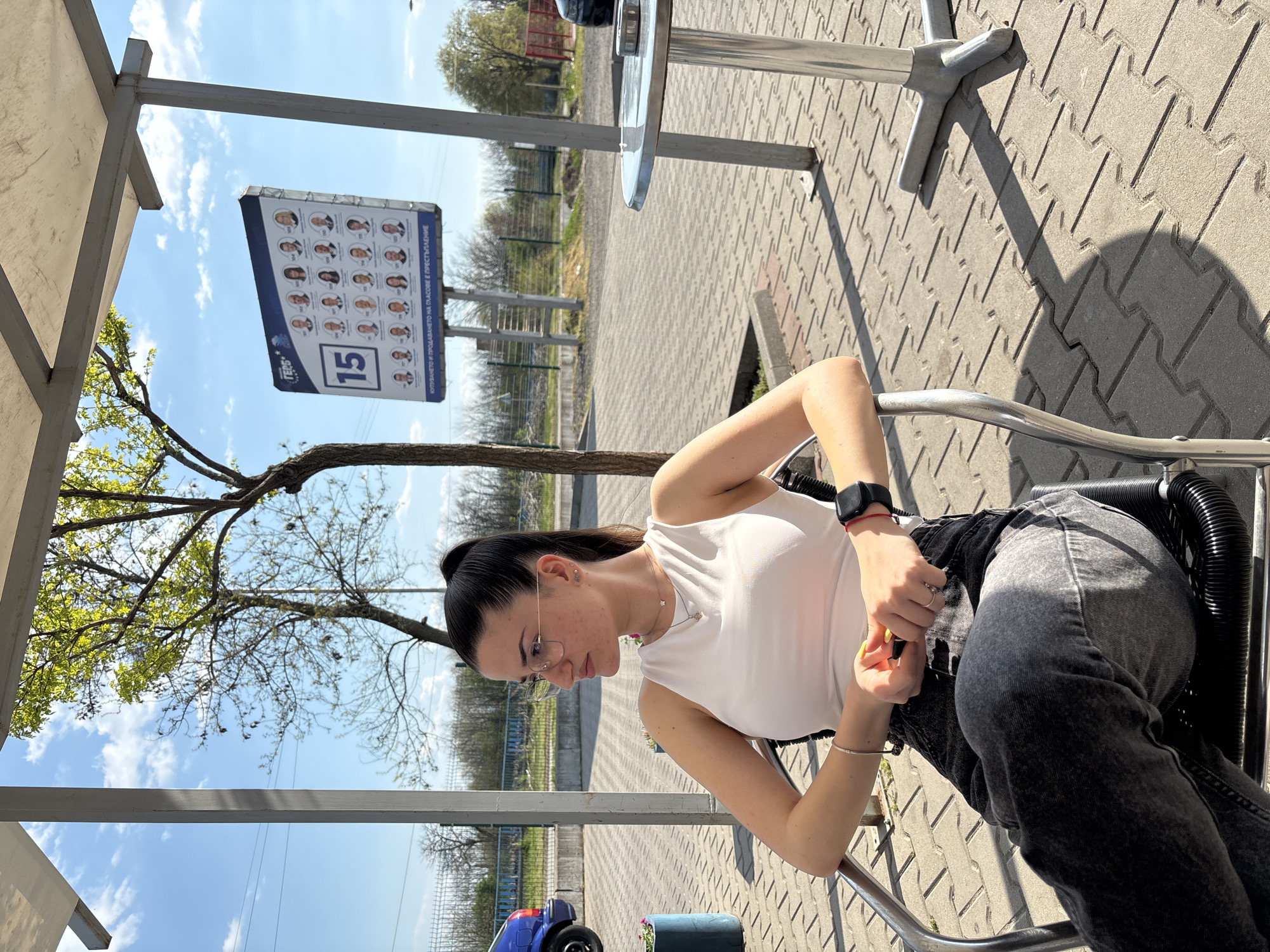 A young woman is seated on a chair at an outdoor café in Chirpan, Bulgaria. She is wearing a white sleeveless top, grey jeans, glasses, and a watch on her left wrist. Her hair is pulled back in a ponytail. The café appears to be situated along a street with trees visible in the background. There is a political sign with photos of candidates posted nearby, indicating upcoming elections. The overall scene suggests a casual, public setting on a sunny day.