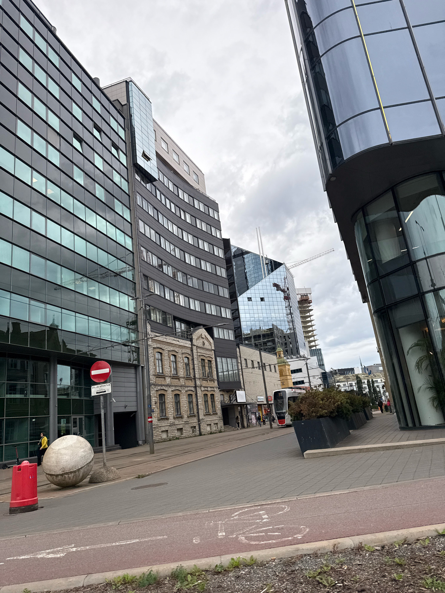 In Tallinn, Estonia, a modern cityscape unfolds with a blend of contemporary architecture and older structures. The foreground features a paved street with red and gray sections, a red bollard, and a large, spherical concrete sculpture. A traffic sign indicates "VALJA ARVATUD," meaning "OUTSIDE THE SCOPE" or "EXCEPTED."
Dominating the left side is a sleek, multi-story building with a dark facade and numerous windows reflecting the overcast sky. Adjacent to it stands a narrower, older stone building with arched windows. In the mid-ground, a modern tram passes by, accompanied by dark planters and several pedestrians. Further back, a construction site reveals cranes and partially built skyscrapers, showcasing ongoing development. The scene is characterized by a cloudy sky and an urban environment.
