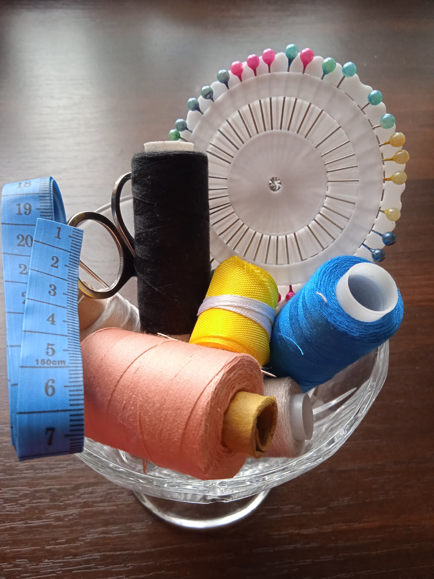 The image depicts a close-up, overhead shot of sewing supplies arranged in a clear glass bowl on a wooden surface. The bowl contains several spools of thread in various colors, including peach, black, yellow, and blue. A blue measuring tape is draped over the side of the bowl, with markings visible up to 7 inches and 19 centimeters. A pair of small scissors is also partially visible within the bowl, along with a needle. To the right of the spools is a white circular pin cushion with numerous colorful pins. The background is a dark brown wooden surface, likely a tabletop. There are no people or animals present in the image, and no discernible action is taking place. The scene appears to be a still life composition of sewing materials. The location is unknown.