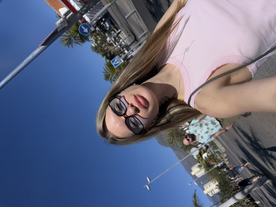 A woman in a pink shirt and glasses takes a selfie outdoors. A street scene unfolds behind her under a clear blue sky. A palm-lined street with buildings and people can be seen. A person wearing a Hawaiian shirt is visible further back. A street pole and traffic signs are also present.