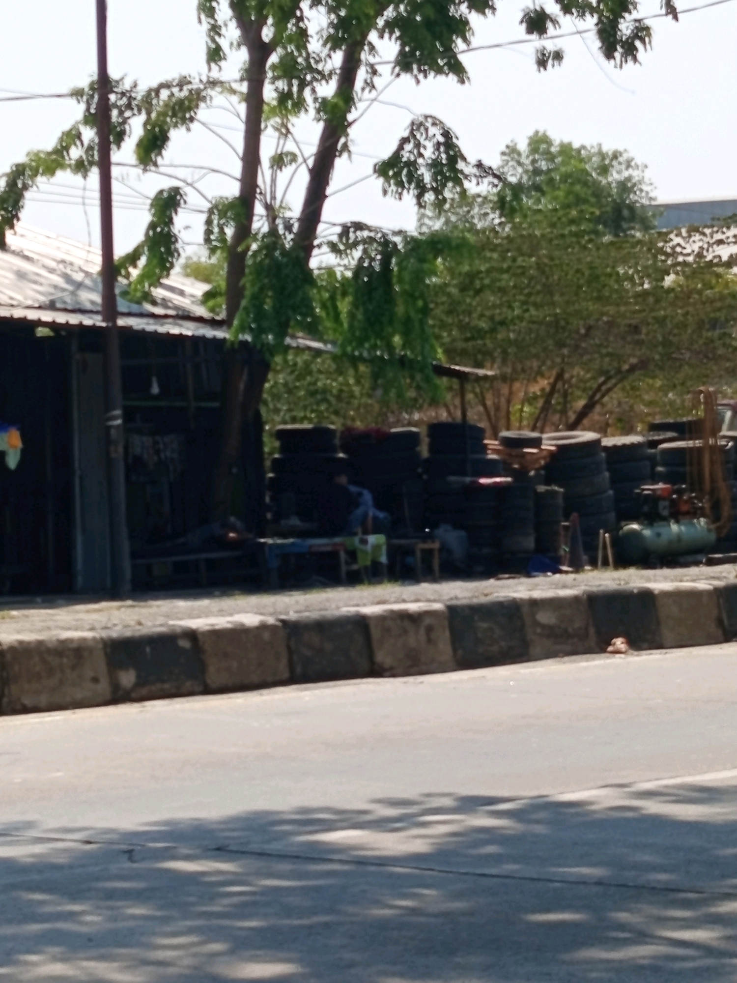 The image depicts an outdoor scene in Karawang, Indonesia, likely a tire repair shop or storage area situated near a road. In the foreground, a paved road is visible with a concrete divider adorned with black and white stripes. Beyond the divider, a collection of stacked tires dominates the mid-ground, arranged in various sizes and quantities. To the left, a small, open-fronted building or stall with a corrugated metal roof and dark walls is present. Inside this structure, it appears that items are stored, and a figure is lying down. Several individuals are observed in the area, with one person seated on a bench and another possibly lying down within the building. Further to the right, a green air compressor unit with an orange engine is situated next to the tire stacks. Trees and foliage are visible in the background, suggesting a semi-urban or roadside environment. The lighting indicates it is daytime, with shadows cast by the trees on the road.