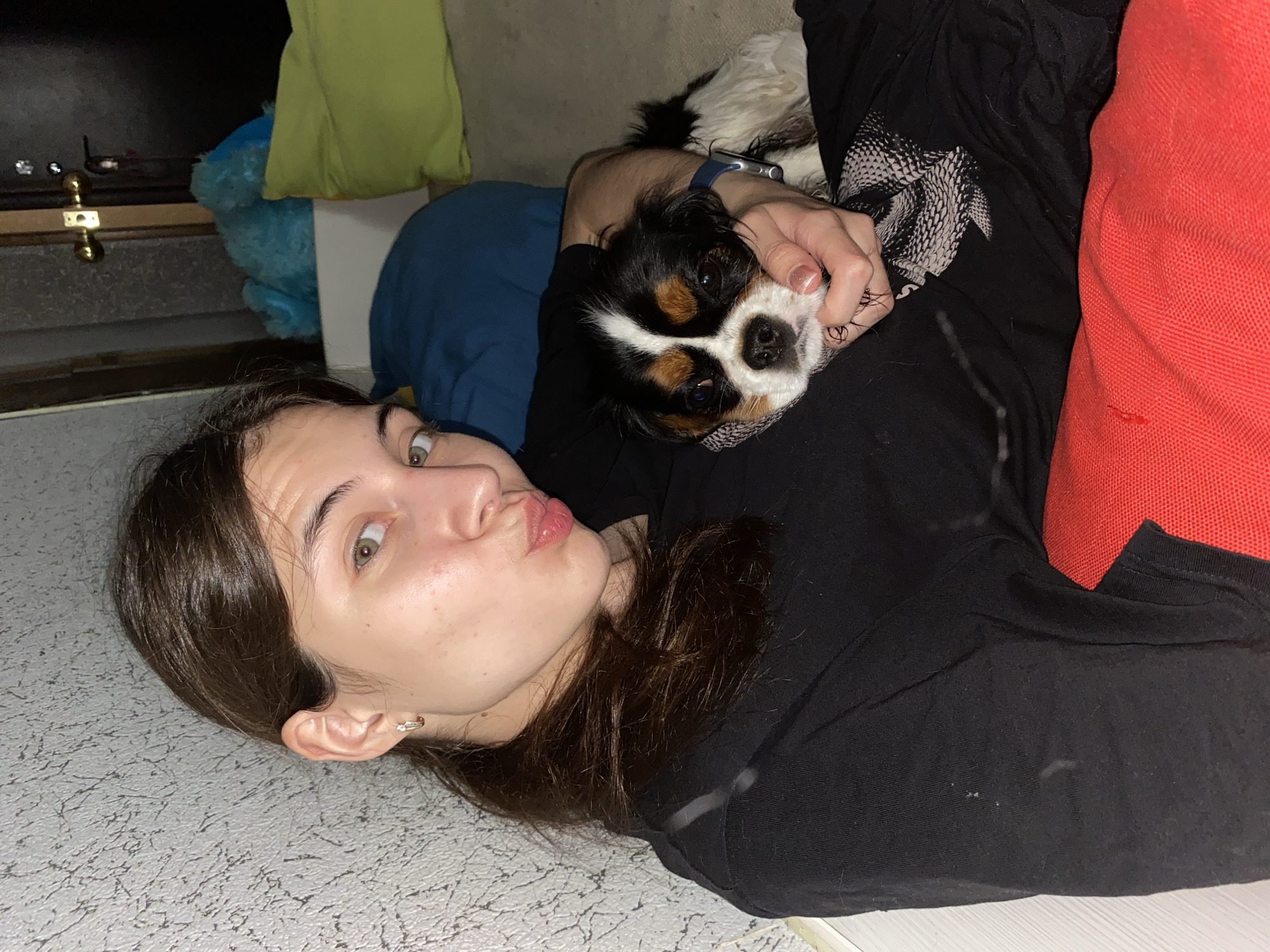 In a dimly lit indoor setting, possibly a residential interior in Votkinsk, Russia, a young female with dark hair pulled into a ponytail is holding a Cavalier King Charles Spaniel. She is looking towards the camera with a playful expression, puckering her lips. The dog is nestled in her arms, its head resting on her hand. The background reveals a textured wall, a door with brass hardware, and various soft furnishings including blue, green, and red cushions or pillows. The woman is wearing a black t-shirt, and the dog has a distinctive tricolor coat of black, white, and tan.