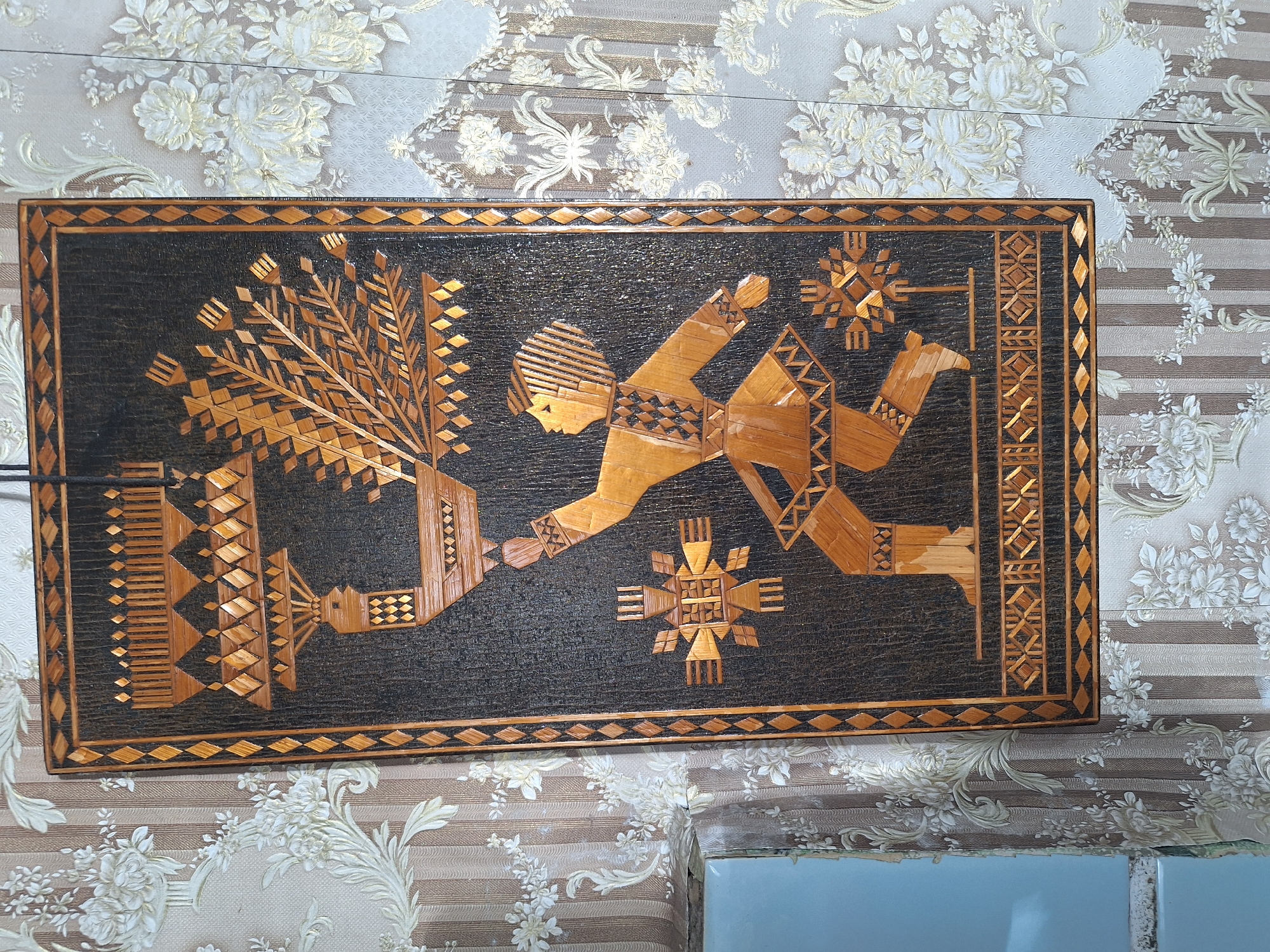The image depicts a piece of folk art, possibly a wall hanging, crafted from straw or a similar natural material. The artwork features a stylized human figure in traditional attire, dancing or presenting a bird. The figure is depicted in motion, with one leg forward and arms outstretched. The bird, also stylized, is perched on the figure's outstretched hand, with a large, elaborate tail. The artwork is framed by a border of diamond and triangular patterns. The background of the artwork is a dark, textured material. The wall behind the artwork is covered in patterned wallpaper featuring vertical stripes and floral motifs. The overall style of the artwork suggests a connection to traditional Slavic or Eastern European crafts. The location is identified as Блеўчыцкі сельскі Савет, Belarus.