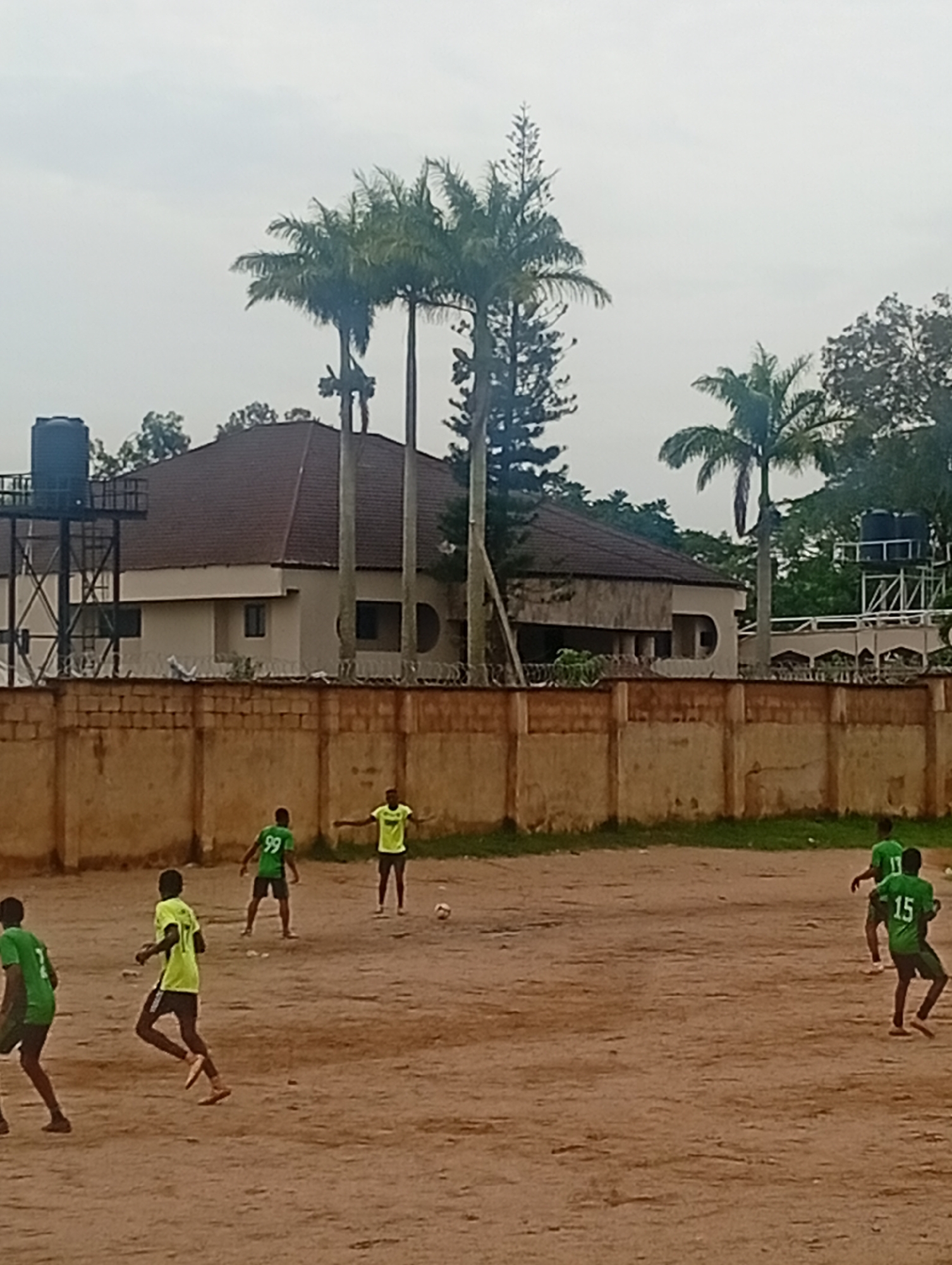 A group of young men are playing soccer on a dirt field in an urban setting, possibly Jos, Nigeria. In the foreground, a player wearing a green jersey with the number 15 on the back is running towards the right of the frame. Another player in a lime green jersey with the number 99 is visible in the middle ground, facing away from the camera. To their right, a player in a bright yellow jersey is facing the camera with his arms outstretched. In the background, a wall separates the field from a building with a brown tiled roof. Palm trees are visible behind the building, and a water tower can be seen on the left side of the image. The sky is overcast.