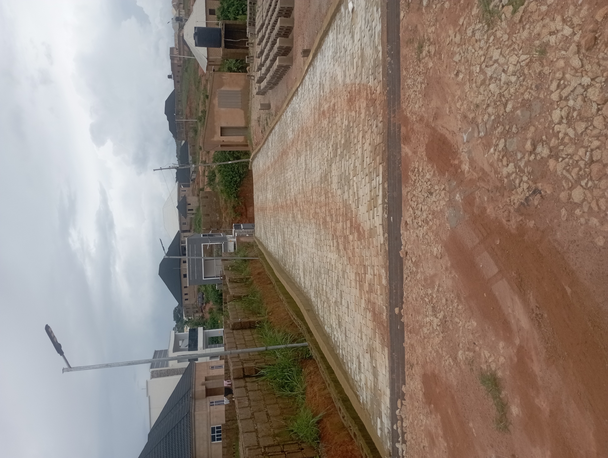 The image depicts a construction site in the city of Rafin Bauna, Nigeria. In the foreground, a dirt path is visible, transitioning into a newly paved driveway made of interlocking concrete blocks. The driveway leads uphill towards a row of construction materials, including stacks of bricks and concrete blocks, and several unfinished buildings in the background. A street lamp stands to the left of the driveway. The sky is overcast, suggesting cloudy weather. The overall scene conveys a sense of ongoing development and construction in a residential area.