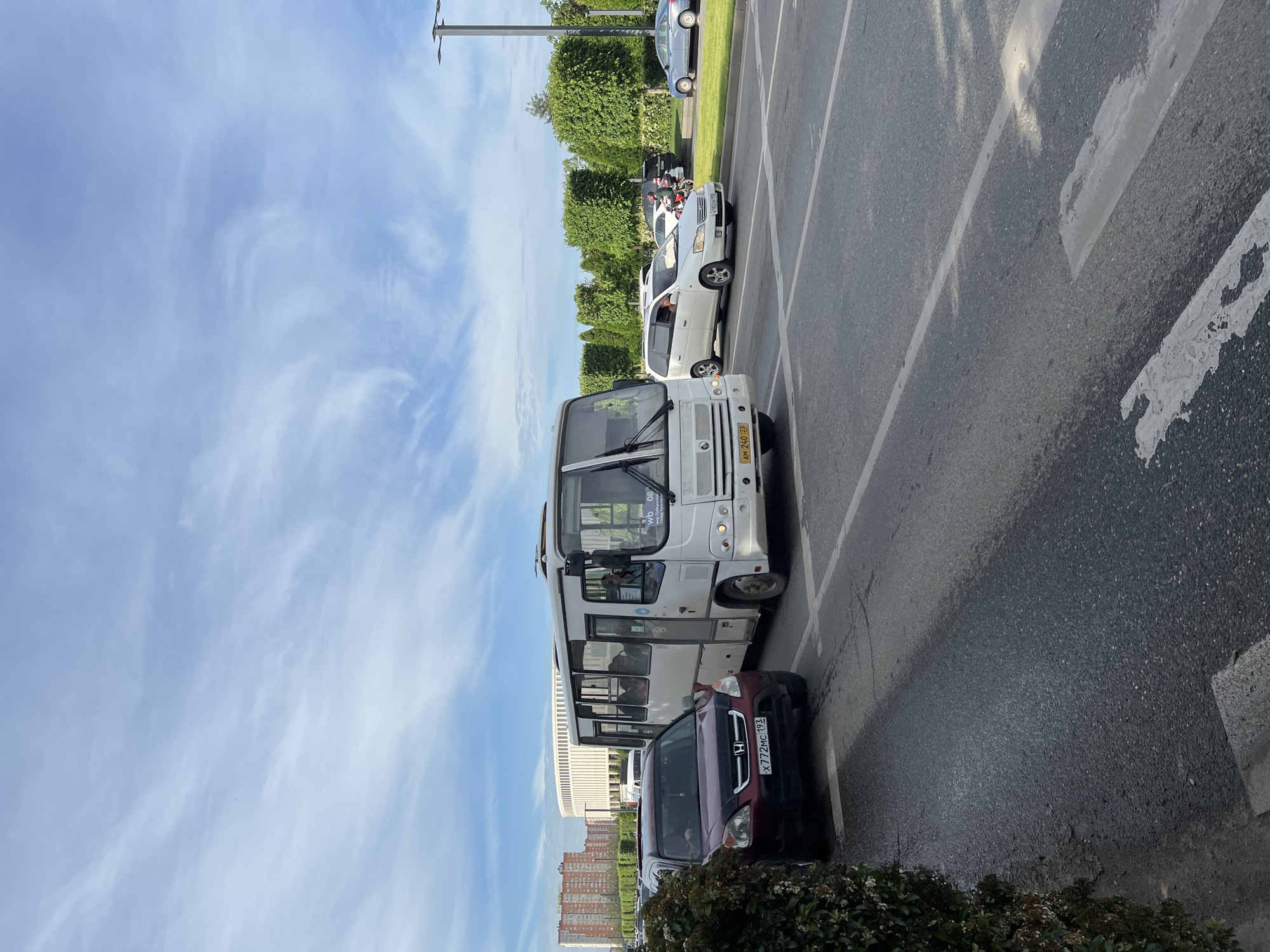 The image depicts a street scene with several vehicles parked along a road in Krasnodar, Russia. A white bus is parked next to a dark red car. Further down the road, a white minivan and another car are visible. On the side of the road, there's a line of trimmed green bushes, followed by buildings and apartments in the background. Above, a blue sky is visible, with sparse clouds. The presence of the "AM 240 23" and "X772MC 193" license plates, indicates the location as Russia.