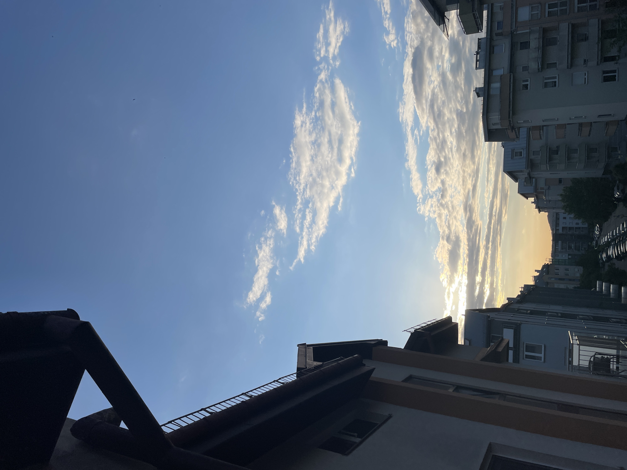 The image captures a view of the sky and buildings in an urban setting. The sky transitions from a clear blue to a brighter, sunset-like glow near the horizon. Clouds are present, particularly in the brighter area of the sky.
Buildings of varying heights line the right side of the frame, their facades partially illuminated by the setting sun. A roof and architectural elements of a lower building are visible in the foreground, casting shadows. No people or specific actions are discernible in the image. The environment is generally indicative of a city, but the specific location is unknown.